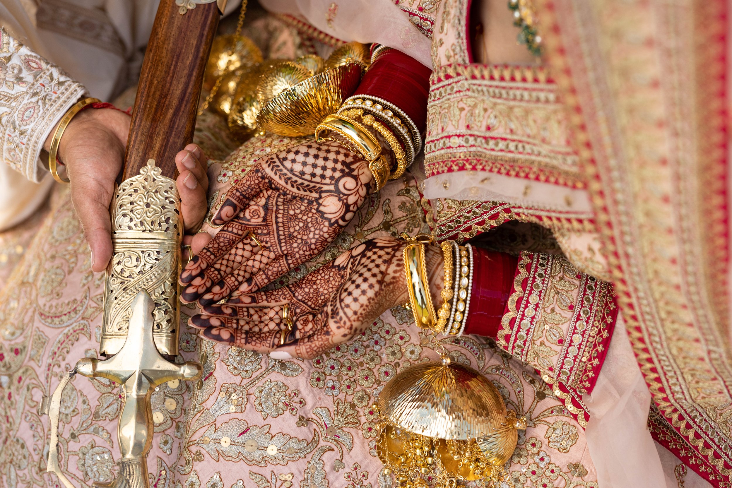 Close-up of traditional Indian wedding attire featuring hands with intricate henna designs, gold jewelry, and colorful embroidered clothing, with a decorative sword and gold ornaments.
