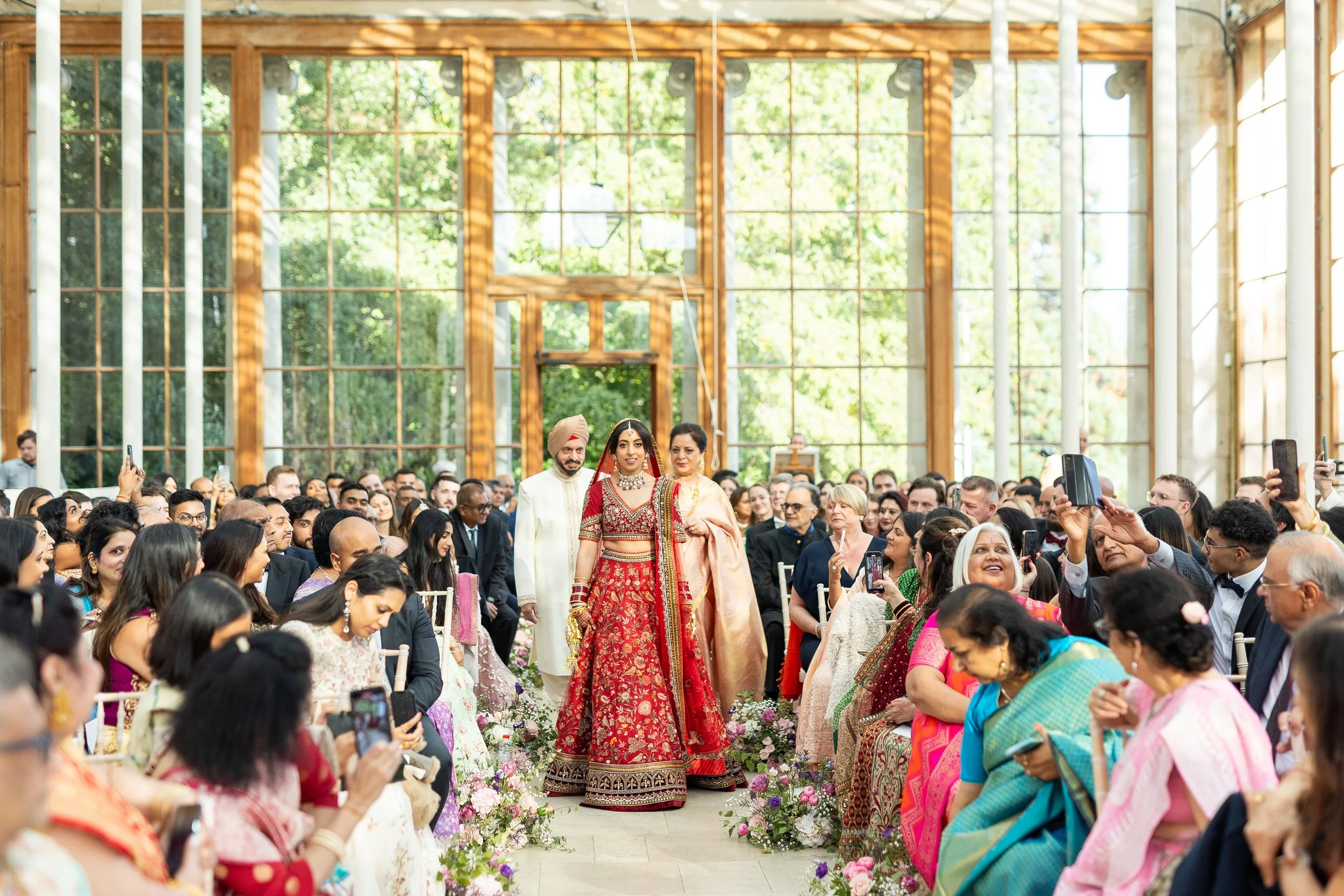A wedding ceremony with a bride in a red traditional Indian dress walking down the aisle, accompanied by a man in a white outfit and a woman in a peach saree, with guests seated on either side taking photos in a glass-walled venue with natural light.