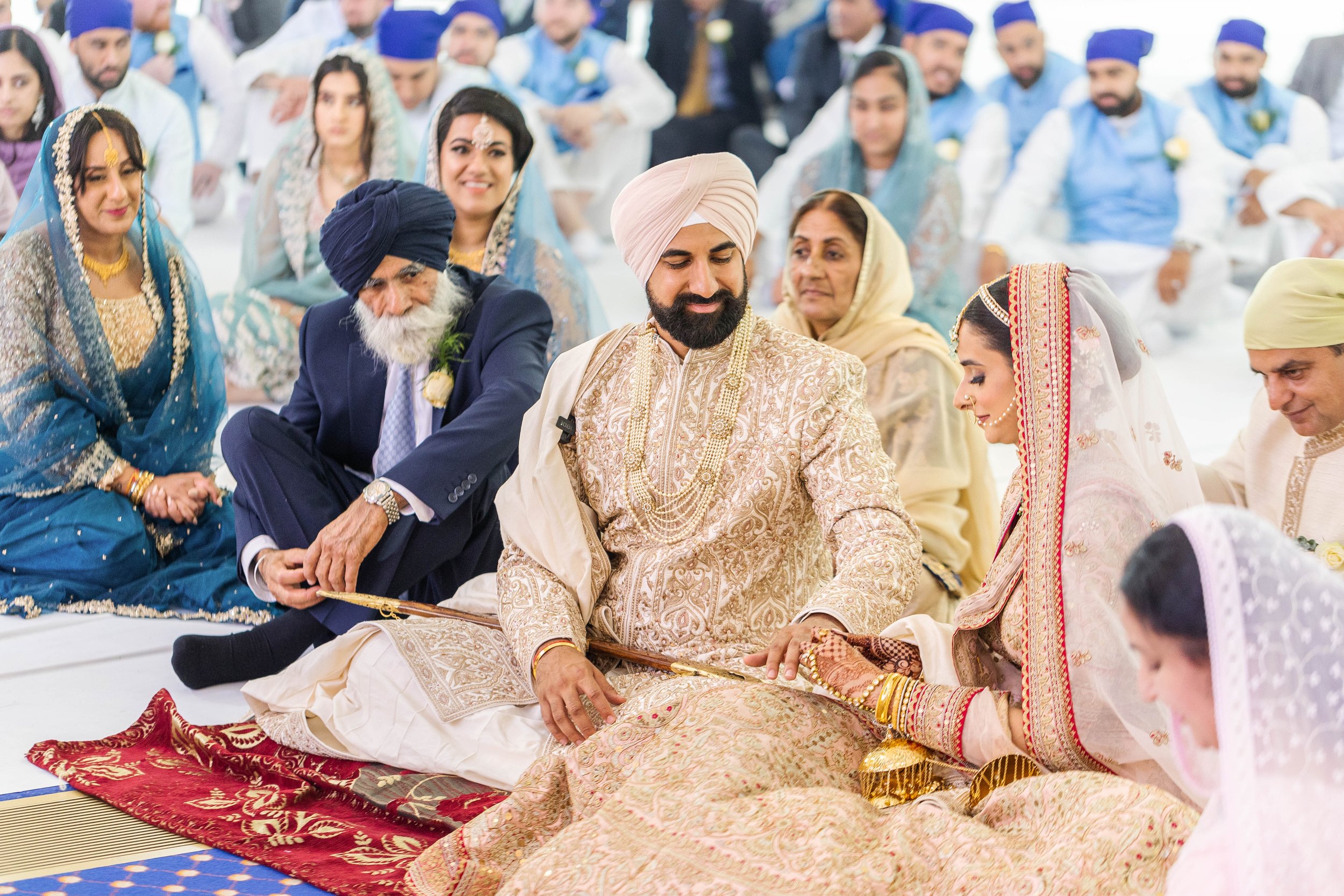 A traditional Indian wedding ceremony with the bride and groom sitting on the floor, dressed in elaborately embroidered outfits, surrounded by family and friends. The bride is wearing a saree with intricate jewelry and henna on her hands, while the g