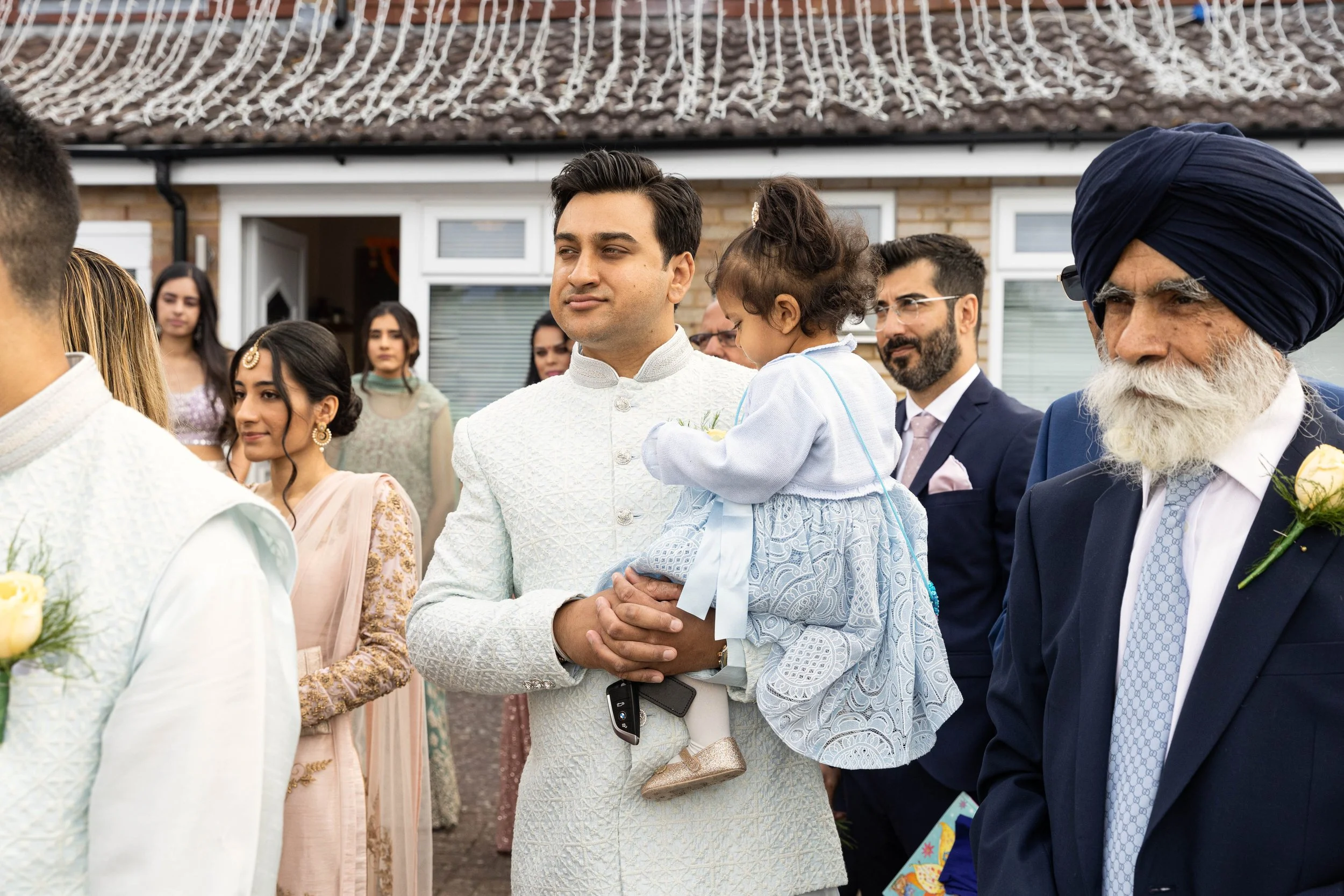 Group of people dressed in traditional Indian and Western attire at a celebration outside, with a man holding a young girl in the foreground.