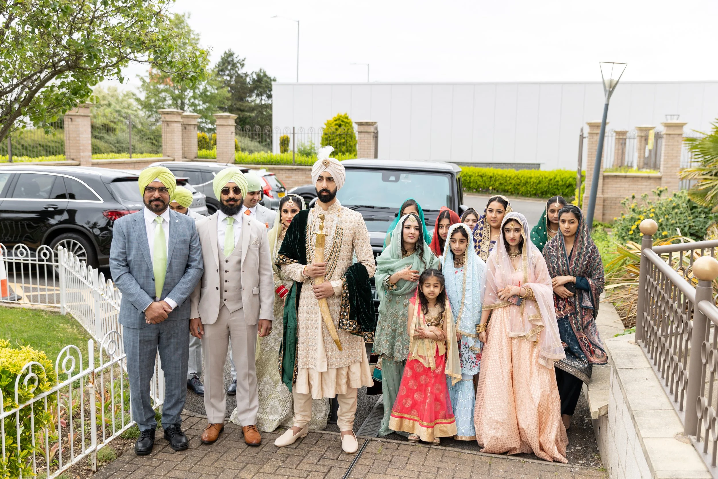 A group of men, women, and children dressed in traditional Indian attire standing outdoors in a parking lot, with cars and greenery in the background.