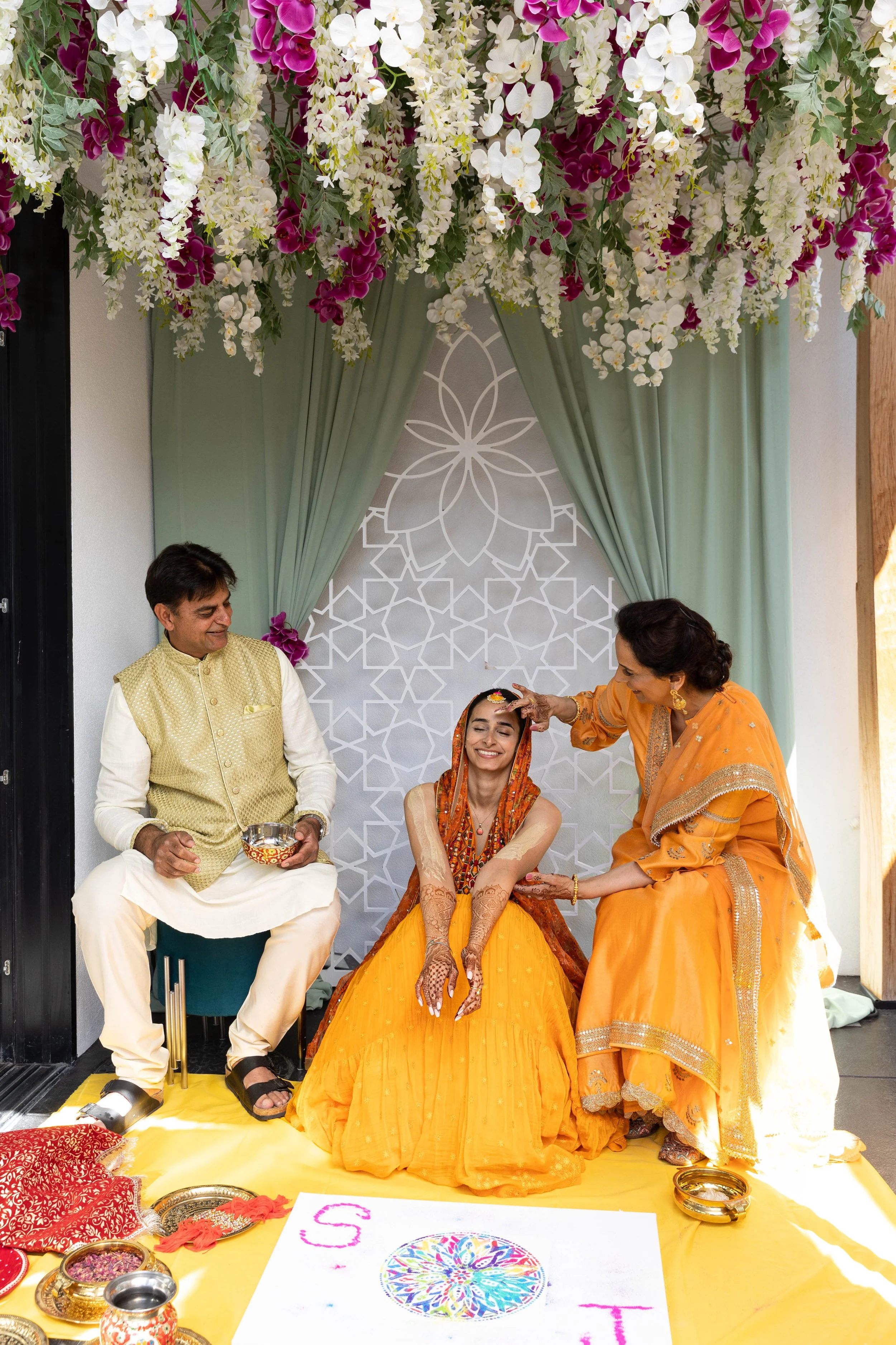 Indian wedding ceremony with a bride sitting on a yellow cloth, surrounded by her parents, during a pre-wedding ritual. The bride is smiling, dressed in a orange outfit, with traditional henna on her hands and decorated with jewelry. An elder woman, 