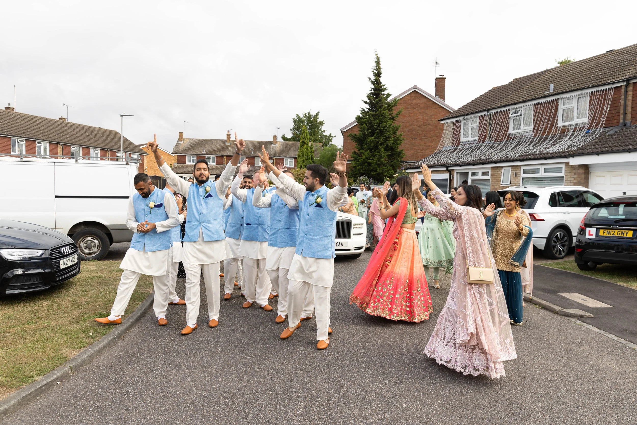 Group of people dancing and celebrating outdoors in a residential area during a cultural or wedding event, with men dressed in traditional South Asian attire and women in colorful, embroidered dresses.