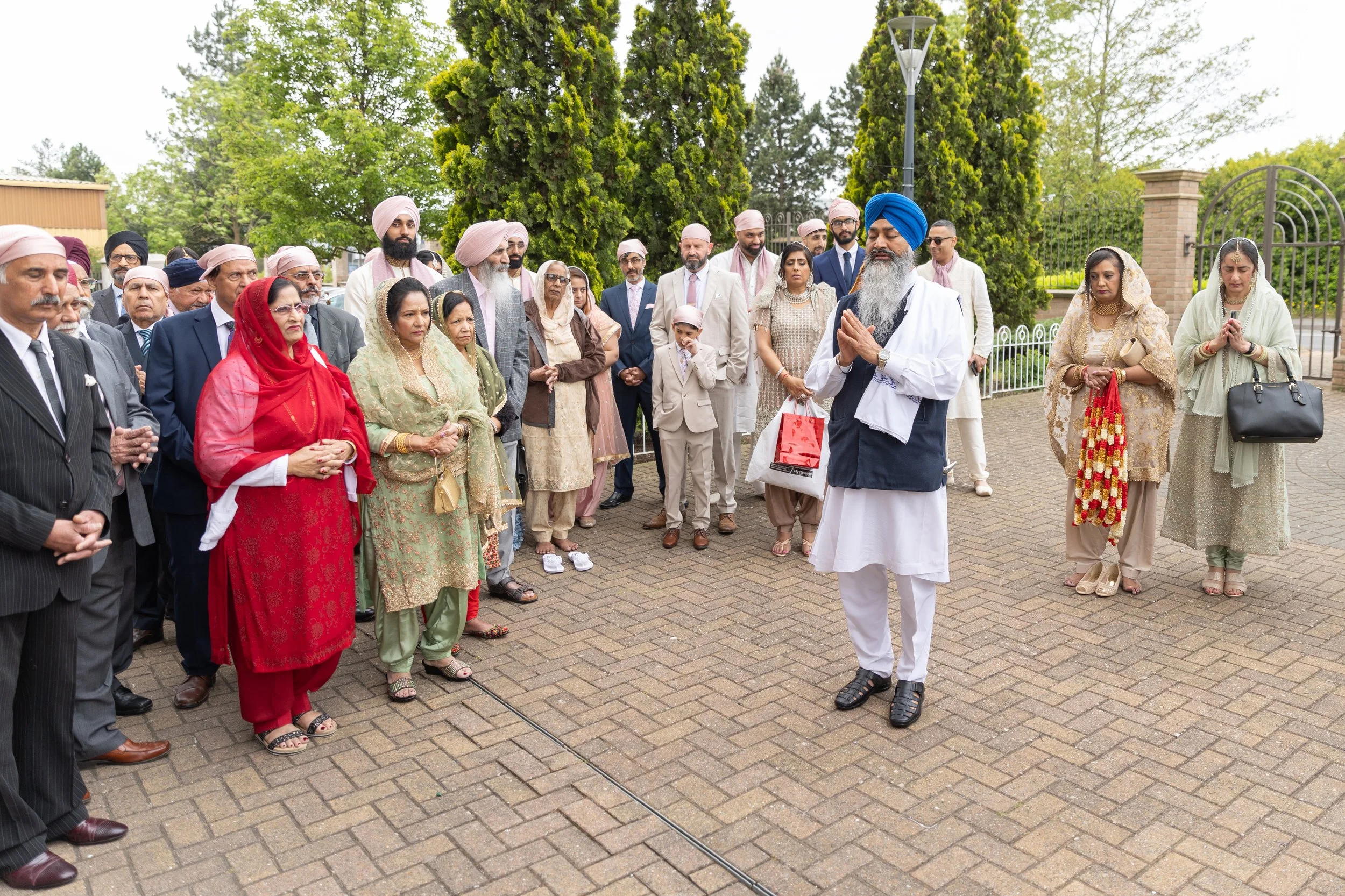 A group of people dressed in traditional and formal attire gathered outdoors for a prayer or ceremony, with some people holding their hands in prayer.
