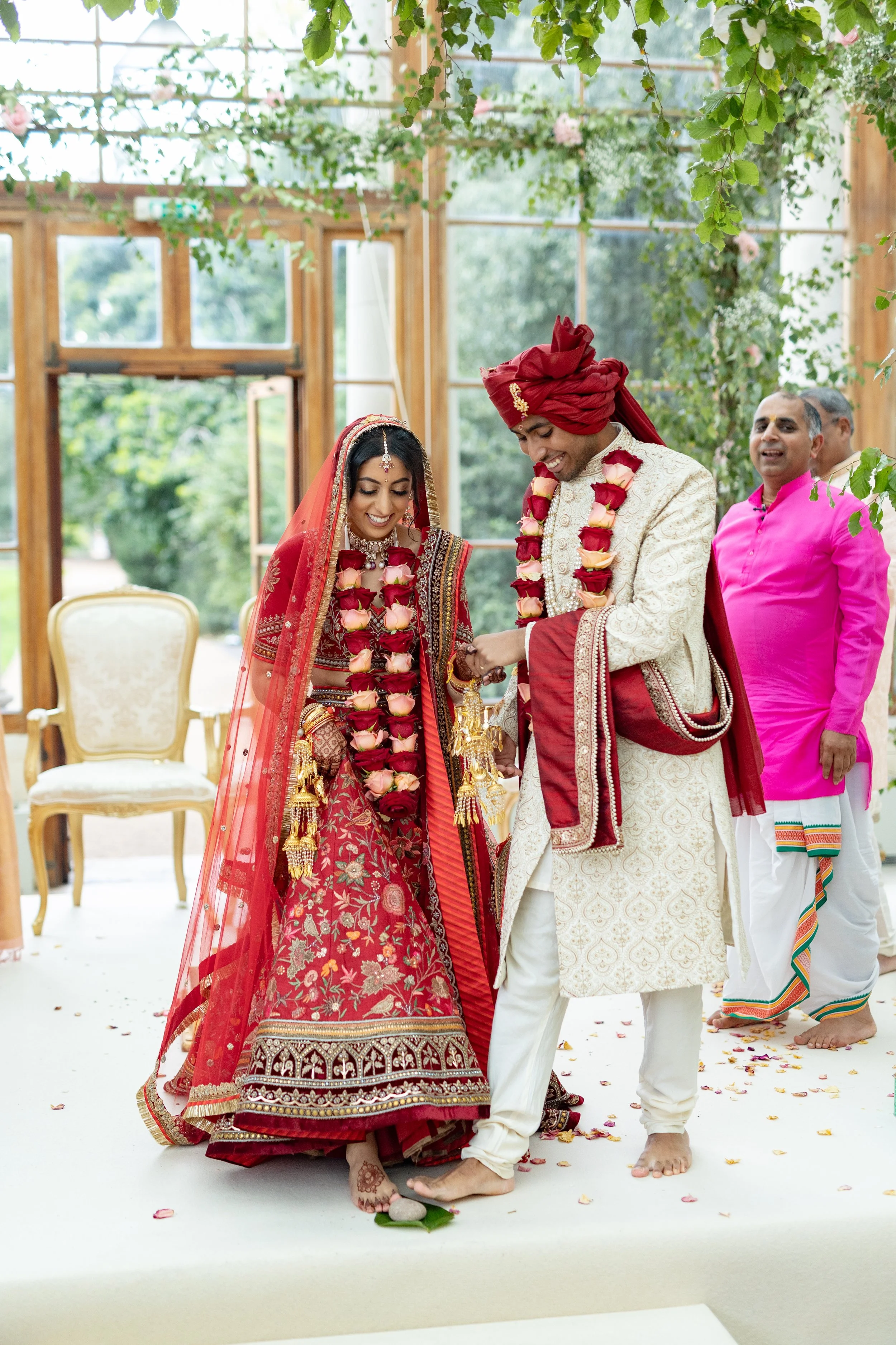 Indian wedding ceremony with bride and groom exchanging rings, wearing traditional clothing and flower garlands, in a decorated indoor setting with greenery.