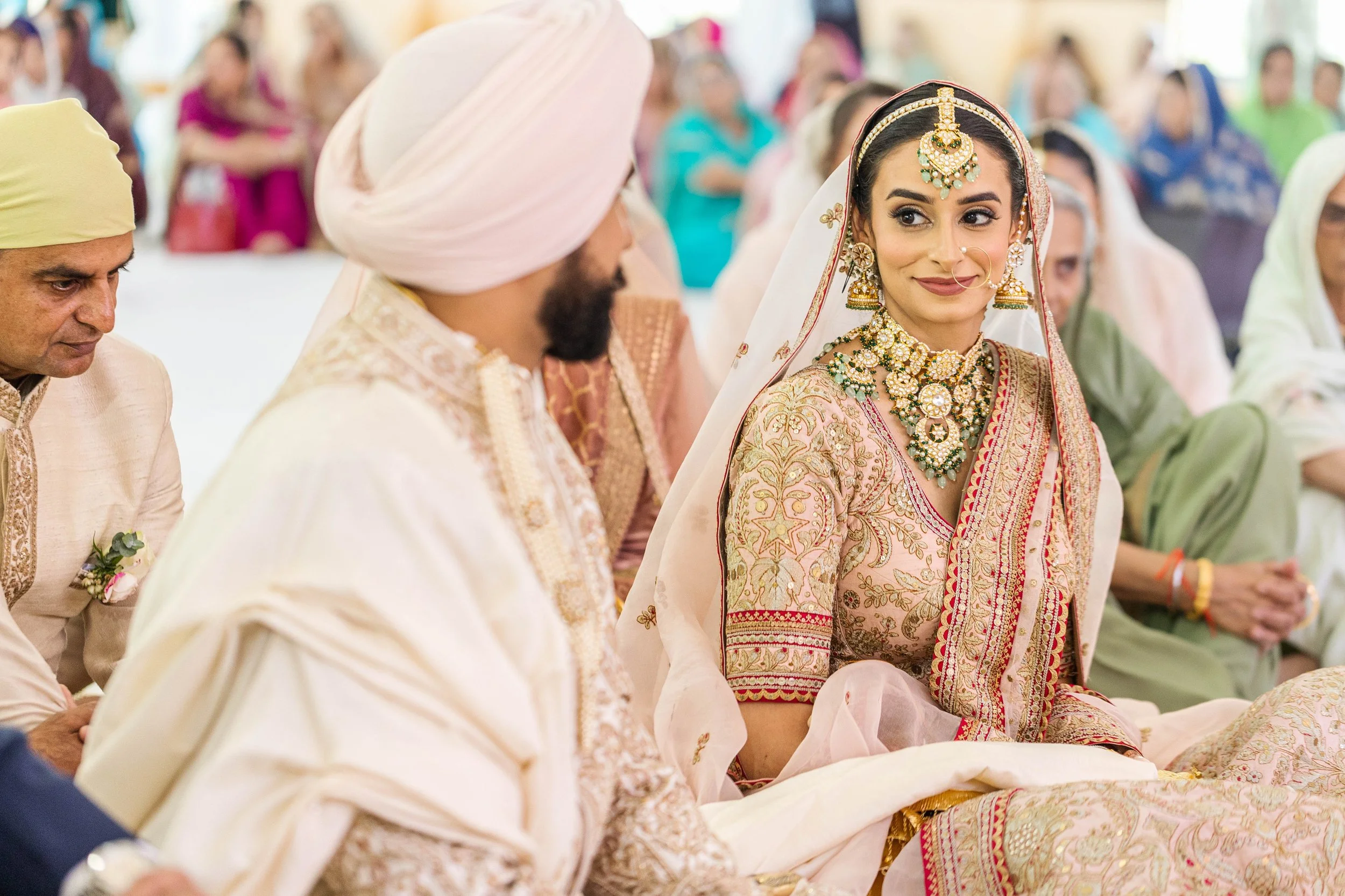 Indian bride in traditional dress and jewelry sitting among guests at a wedding ceremony.