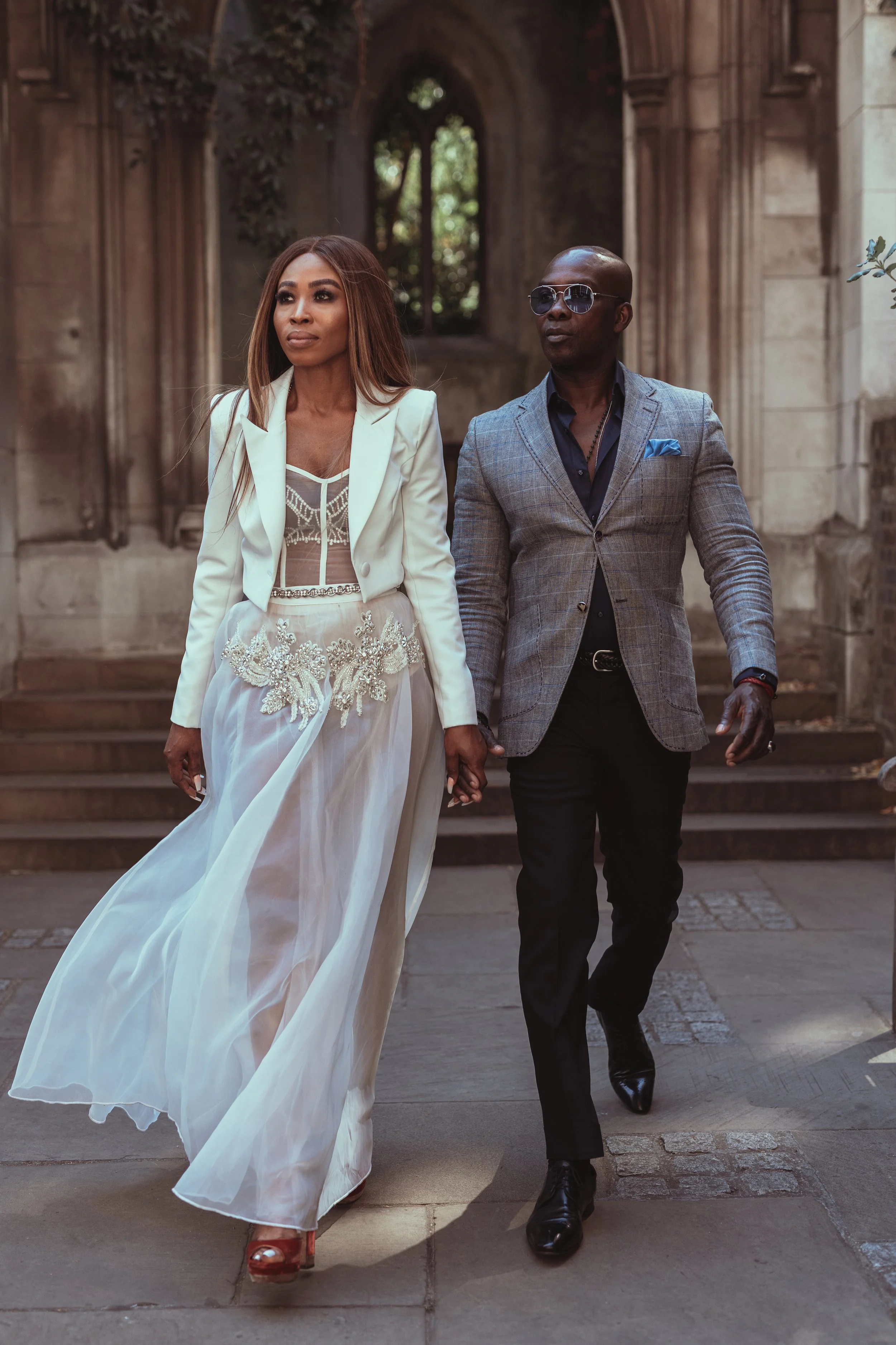 A stylish couple walking hand in hand outside an old stone building, the woman in an elegant white dress and the man in a gray blazer with sunglasses.