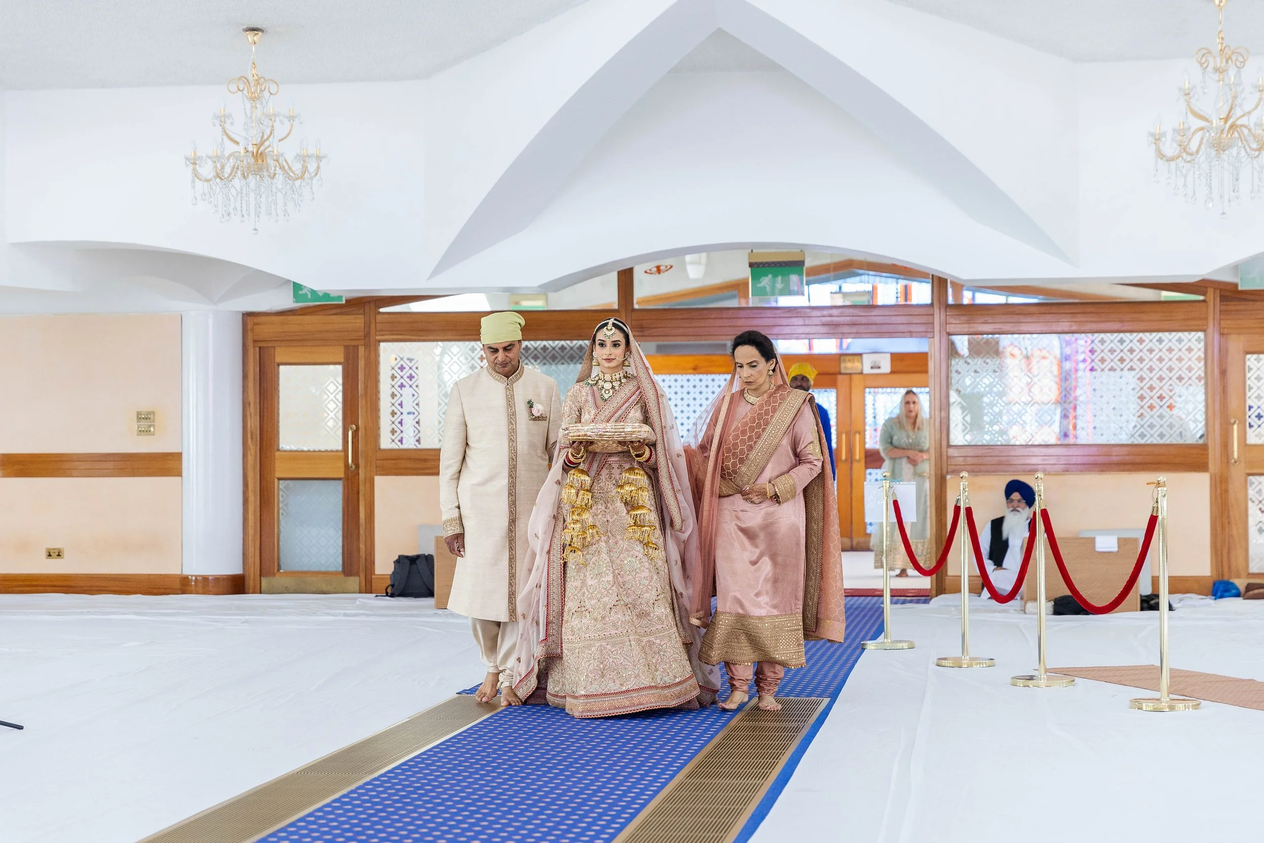 Indian wedding ceremony inside a decorated hall, with the bride dressed in traditional bridal attire and jewelry, walking with her parents along a blue carpet.