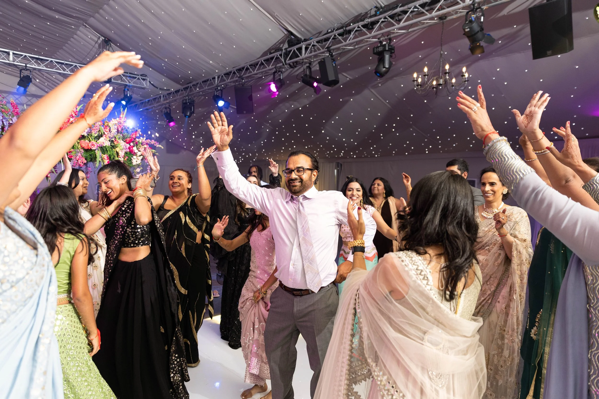 Group of people dancing and celebrating at an indoor event, with a decorated ceiling and colorful lights.