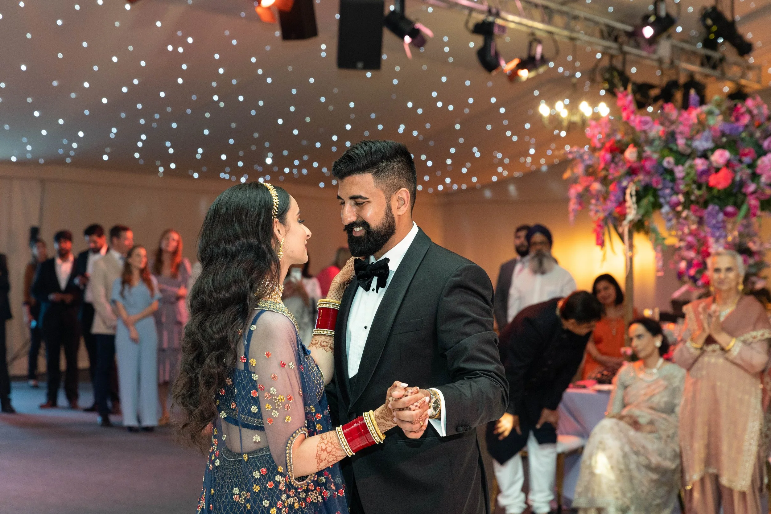 A couple dances at their wedding reception, with the bride dressed in traditional Indian attire and the groom in a tuxedo, surrounded by family and friends in an elegant decorated venue.