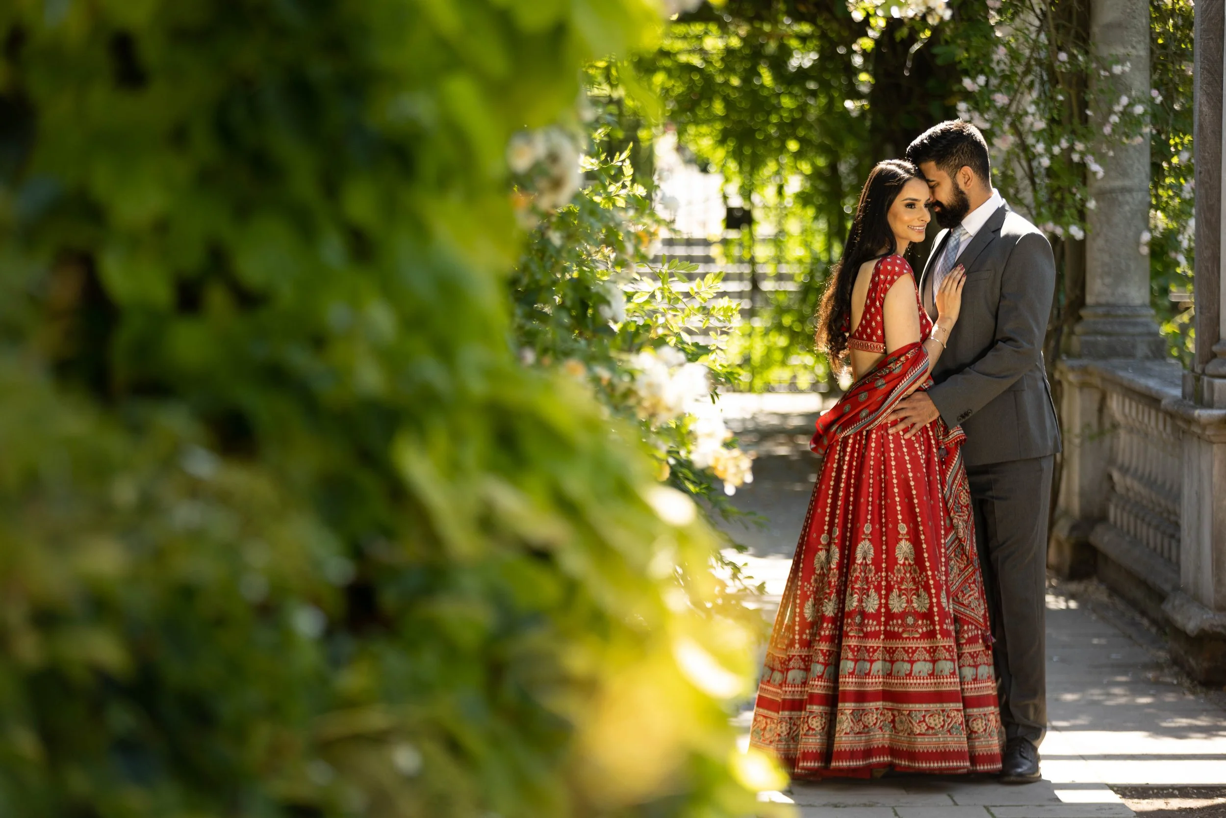 A couple dressed in traditional Indian attire standing close and smiling in a garden or park, surrounded by greenery and leaning against a stone railing.