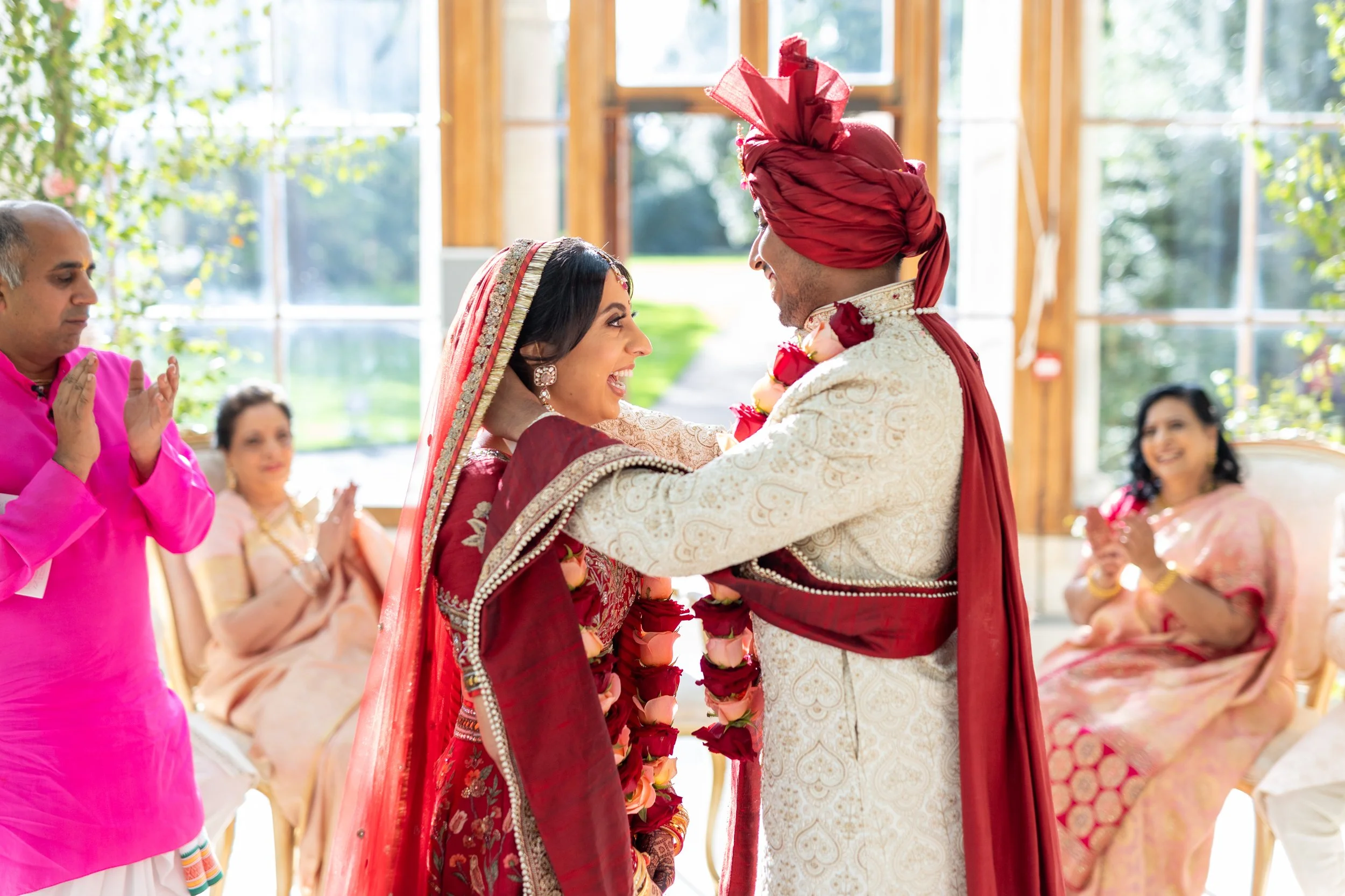 Indian bride and groom in wedding attire smiling and holding each other during a traditional ceremony with family members clapping and celebrating indoors with large windows and greenery outside.