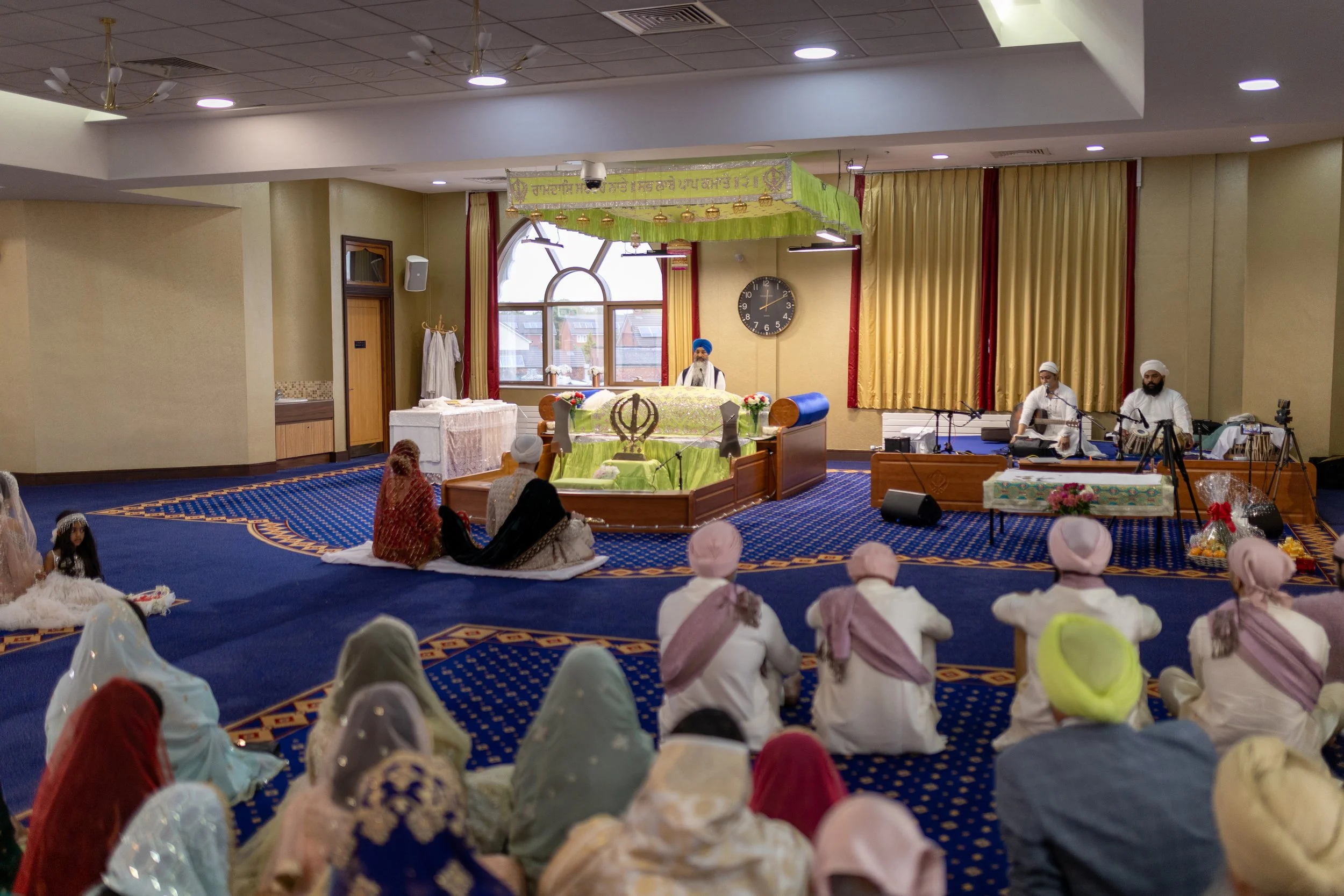 A Sikh religious gathering takes place in an indoor temple, with a Guru Granth Sahib scripture on a raised platform in the center, adorned with cloth and symbols. People are seated on the floor, some wearing turbans and traditional attire, listening 