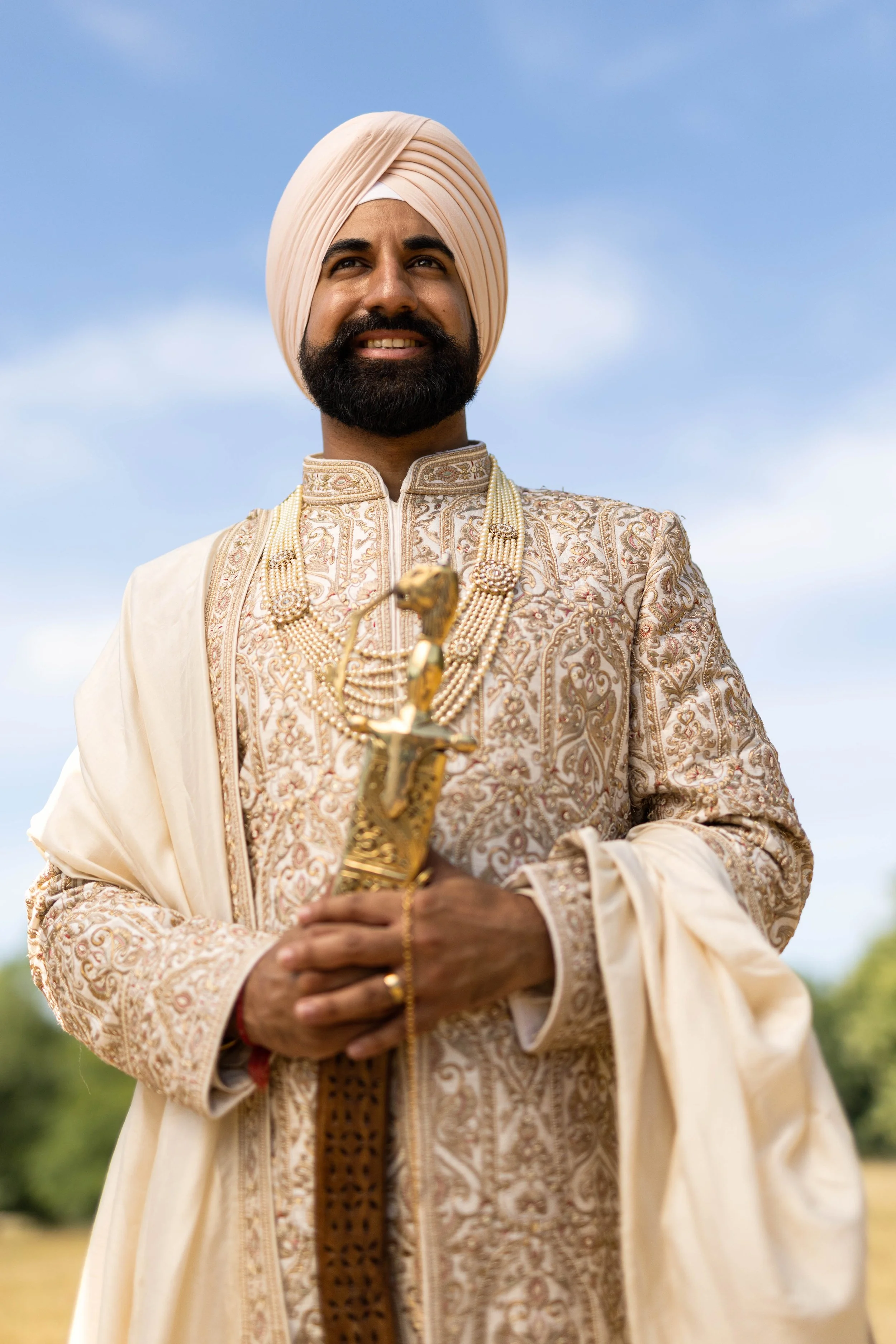 A man wearing traditional Indian wedding attire, including an ornate embroidered sherwani, a beige shawl, and a peach-colored turban, holding a decorative sword, standing outdoors against a blue sky.
