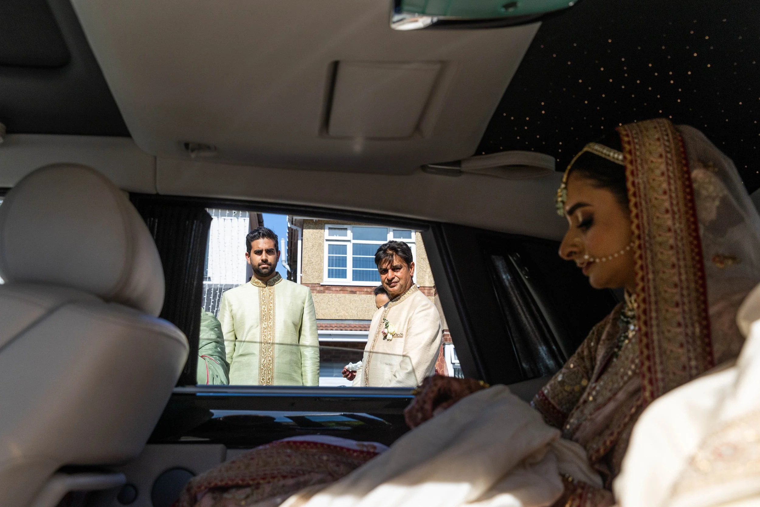 A bride dressed in traditional Indian wedding attire seated inside a vehicle, looking down. Outside the car, three men are dressed in traditional Indian clothing, standing and looking towards the bride.