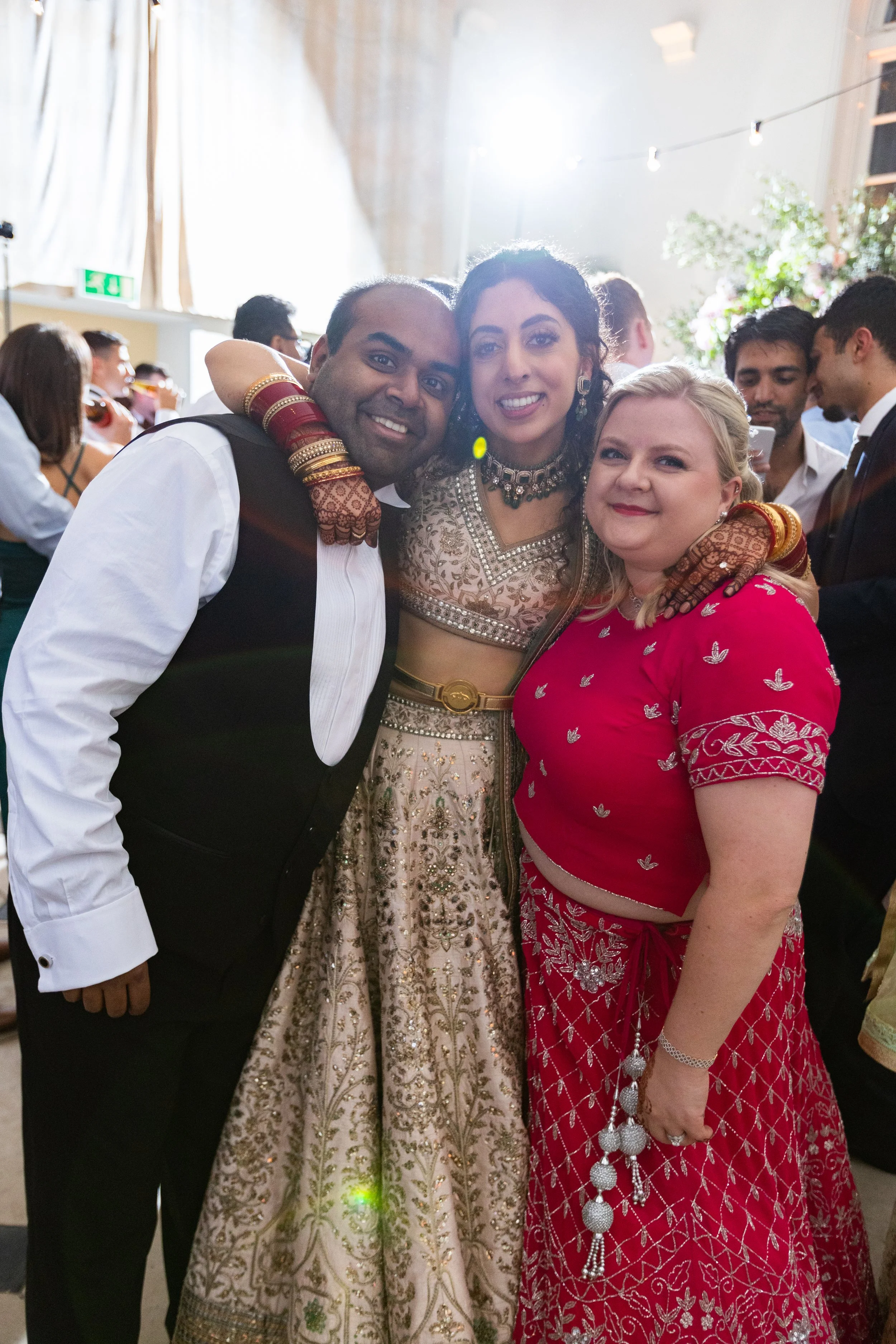 Three people, two women and a man, at a celebration or wedding, dressed in traditional Indian attire, smiling and embracing, with a lively background of other guests and decorations.