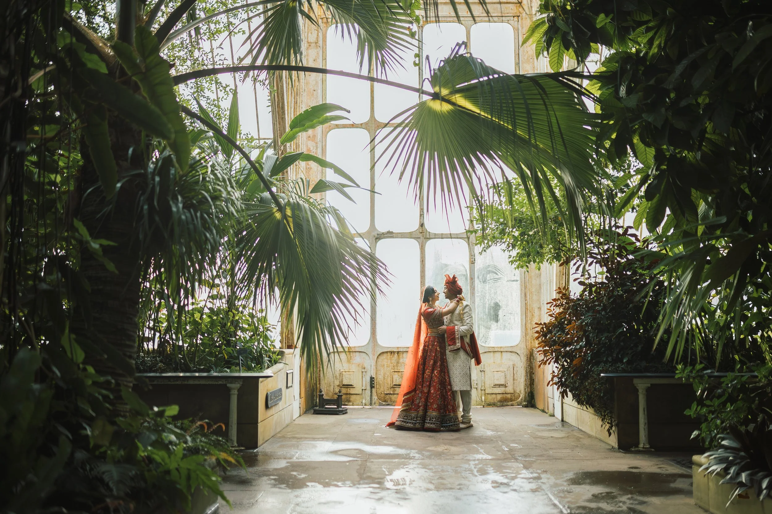 A couple dressed in traditional Indian wedding attire sharing an intimate moment in a lush, indoor greenhouse filled with tropical plants, with a vintage glass door and window in the background.