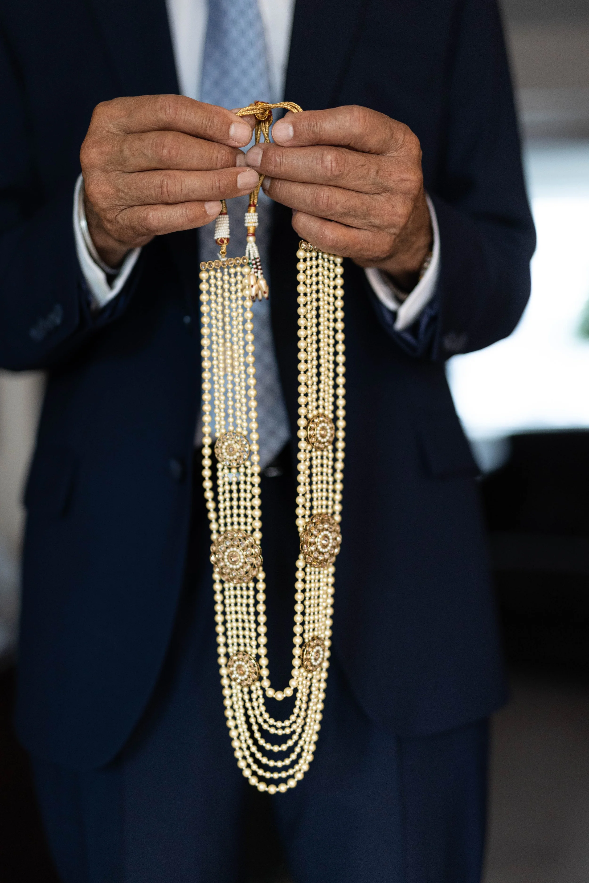 A person in a dark suit holding a multi-strand pearl necklace with ornate gold and pearl pendants.