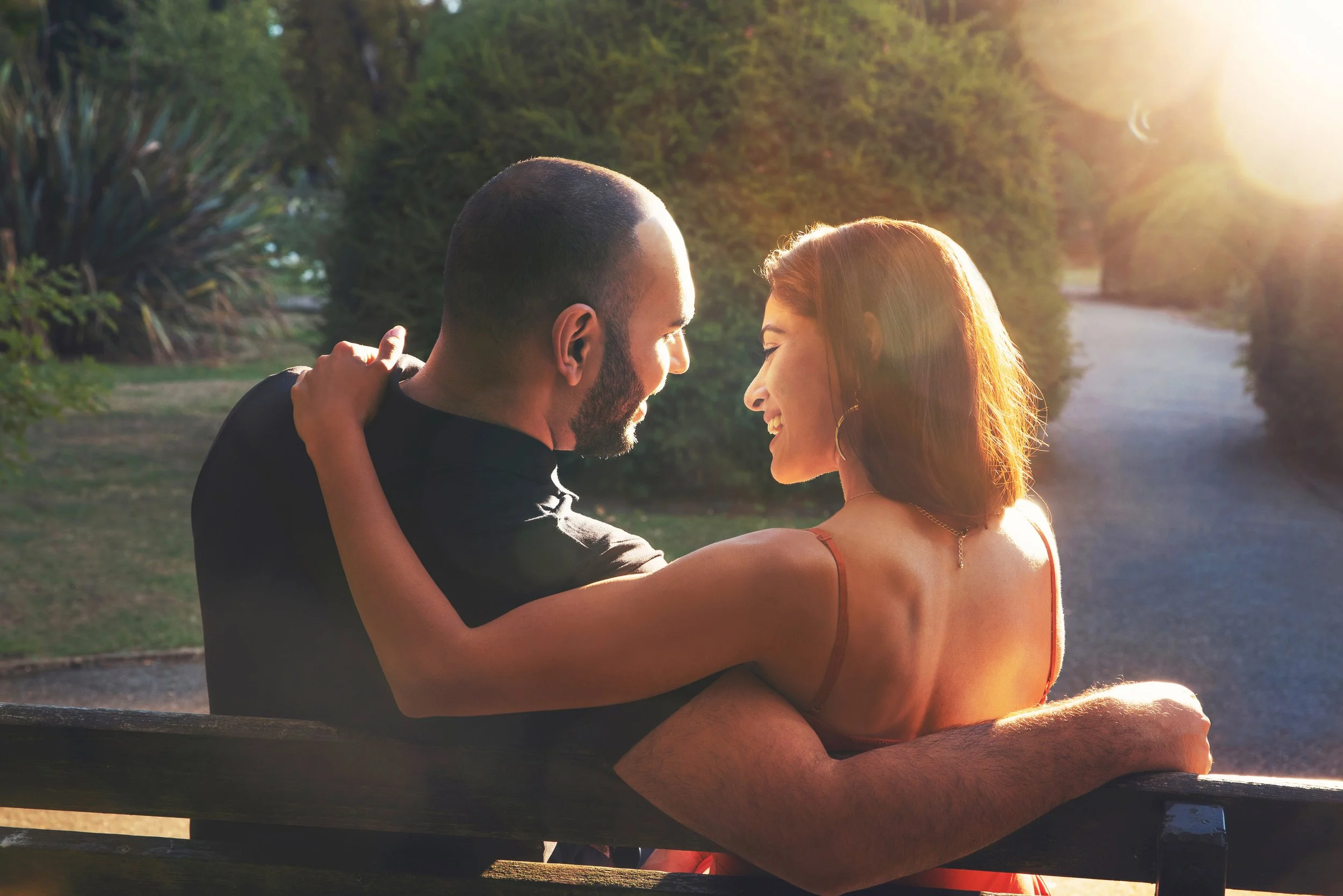 A couple sitting on a park bench, close together and smiling at each other during sunset, with trees and a path in the background.