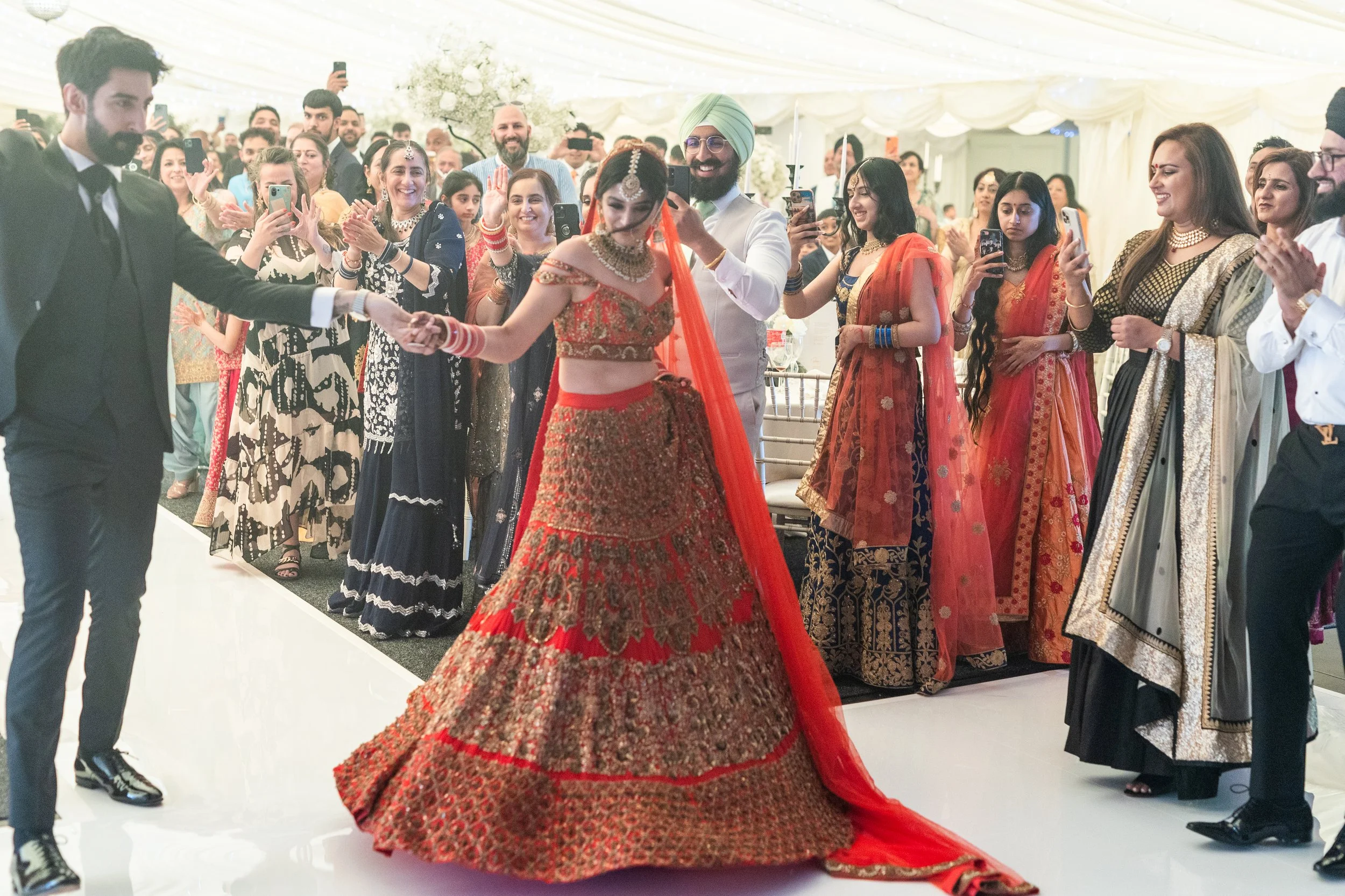 A bride in a red and gold traditional Indian wedding dress dancing with a groom in a white outfit and green turban, surrounded by guests taking photos and clapping inside a decorated tent.