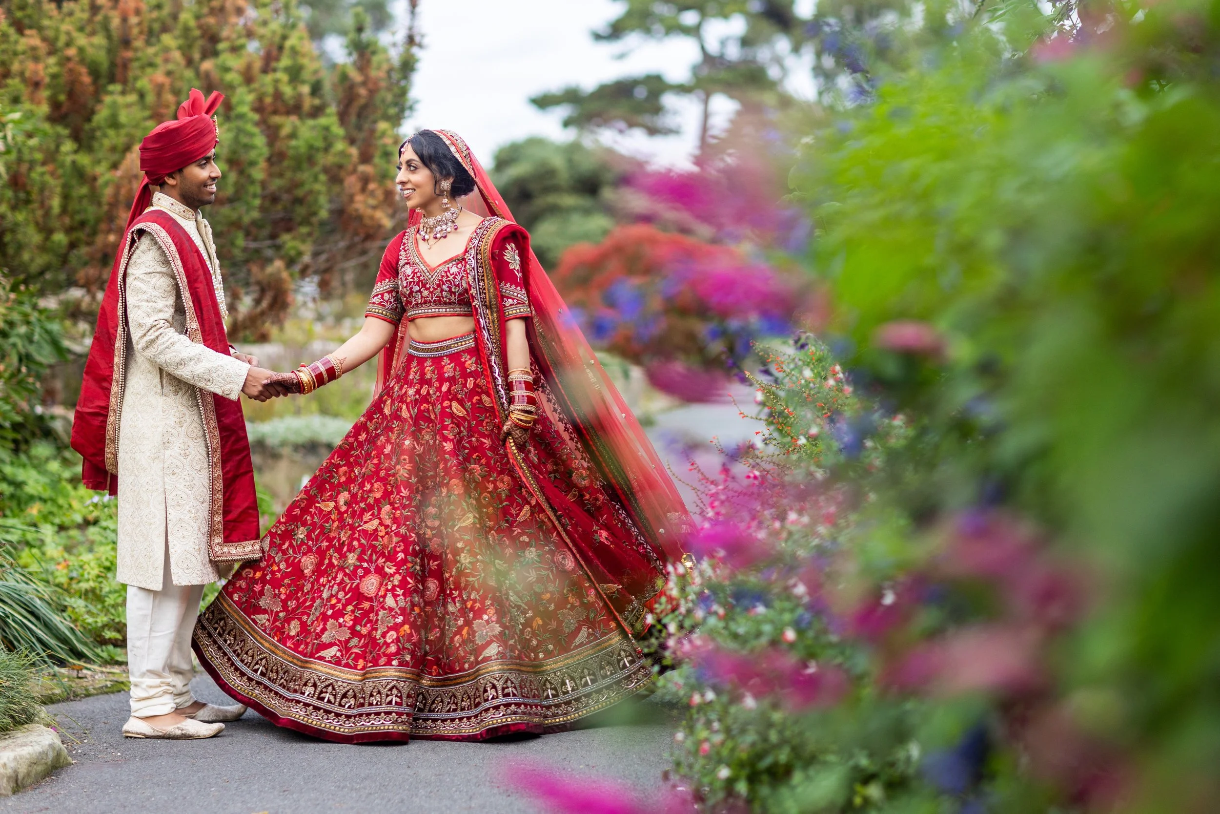 A bride and groom dressed in traditional Indian wedding attire holding hands in a garden with colorful flowers.