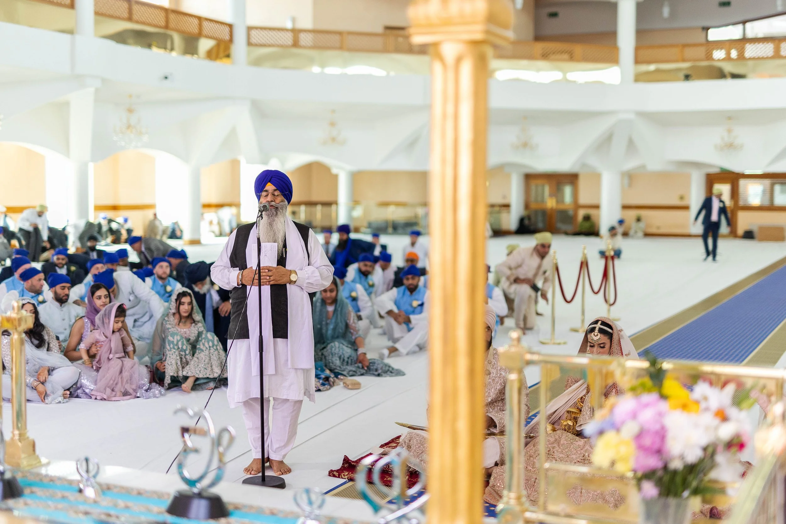A Sikh wedding ceremony taking place indoors with a religious leader speaking into a microphone, surrounded by seated guests dressed in traditional attire, in a spacious hall decorated for the event.