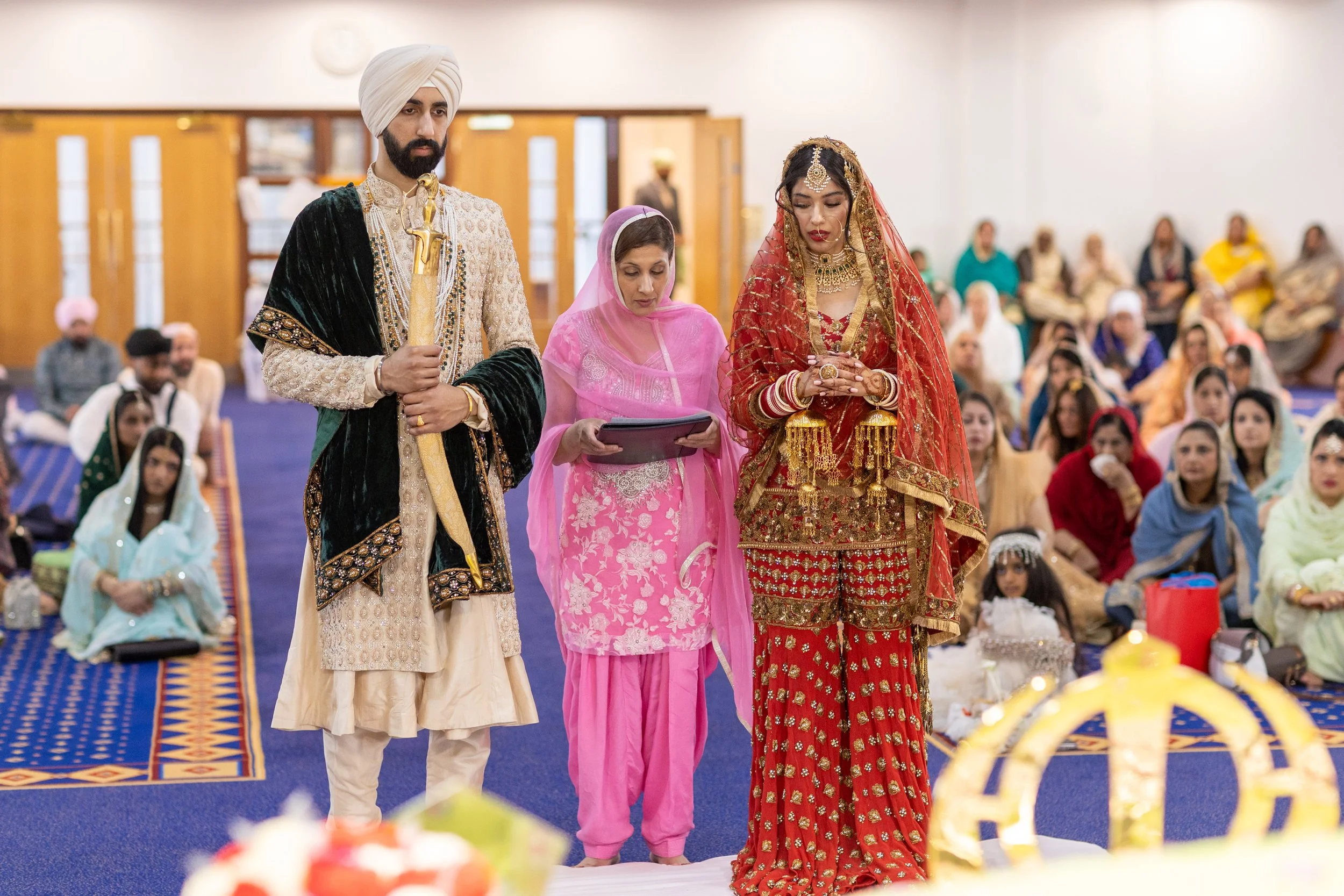 A traditional Sikh wedding ceremony with the bride and groom in ornate attire, officiant, and guests in colorful attire seated on the floor in a decorated indoor hall.