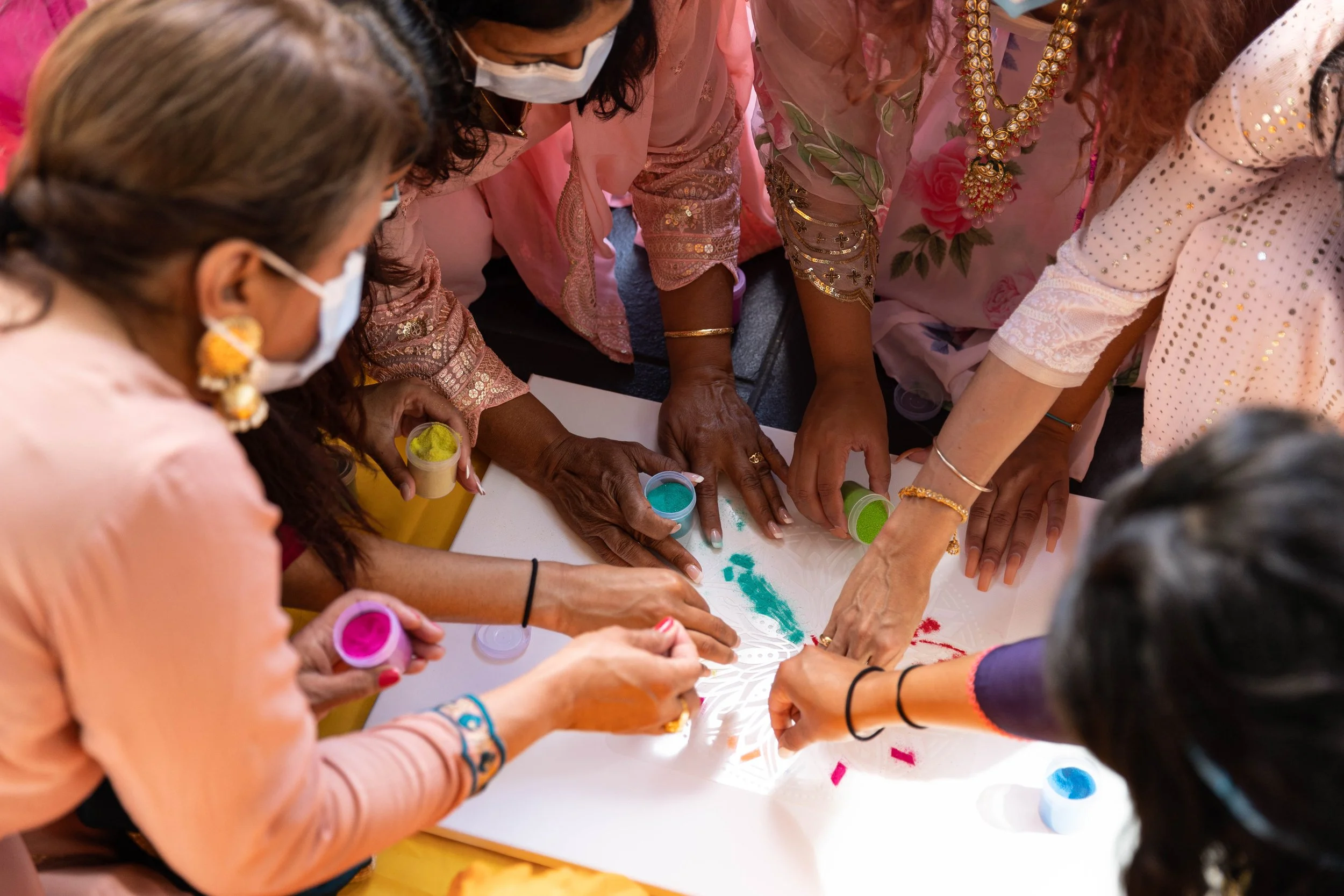 Group of women wearing traditional attire and face masks, participating in a colorful hands-on activity with powders and a paper cutout on a table.