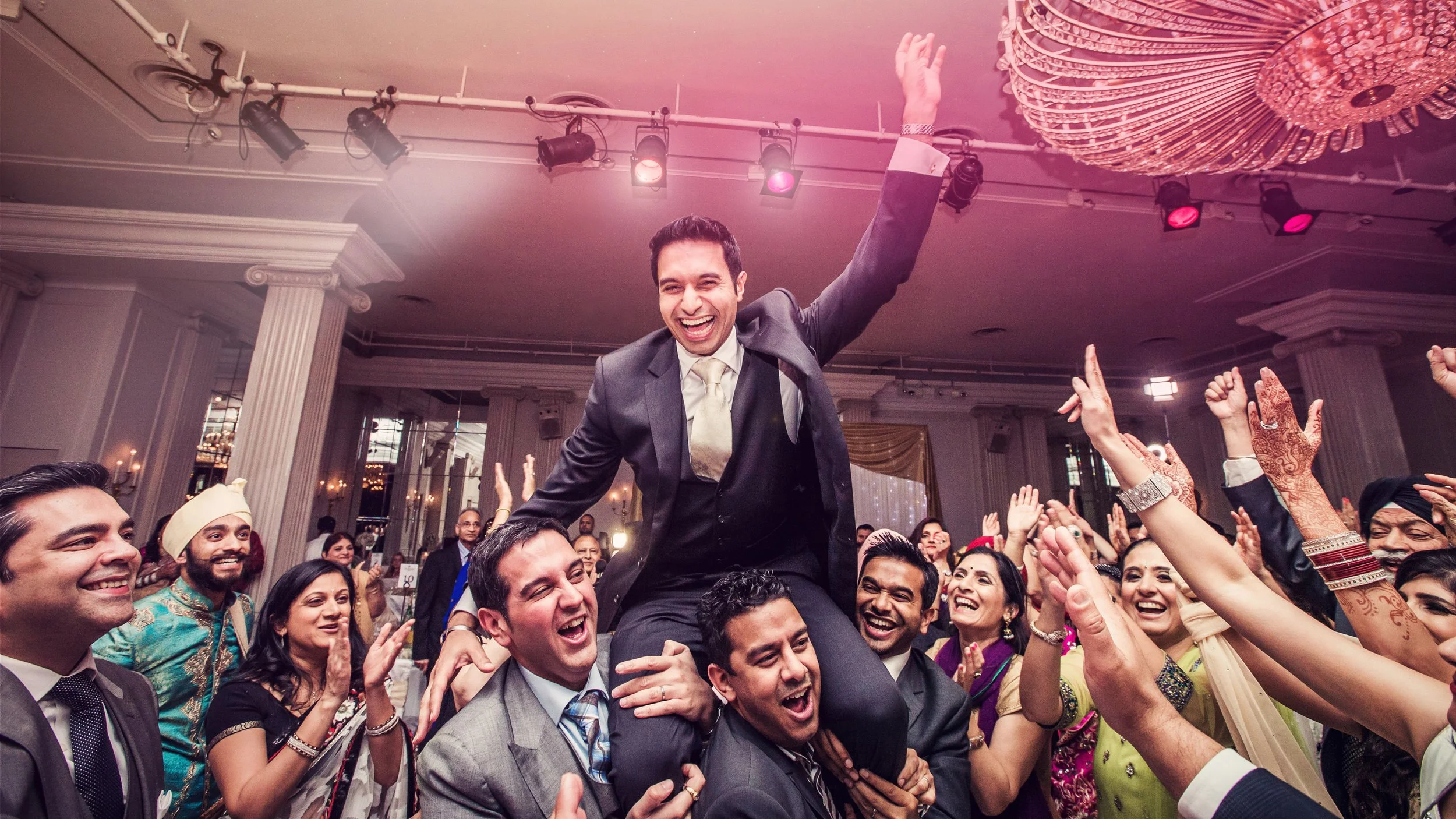 Celebration of a groom being lifted on a chair during a wedding reception, surrounded by joyous guests clapping and smiling.