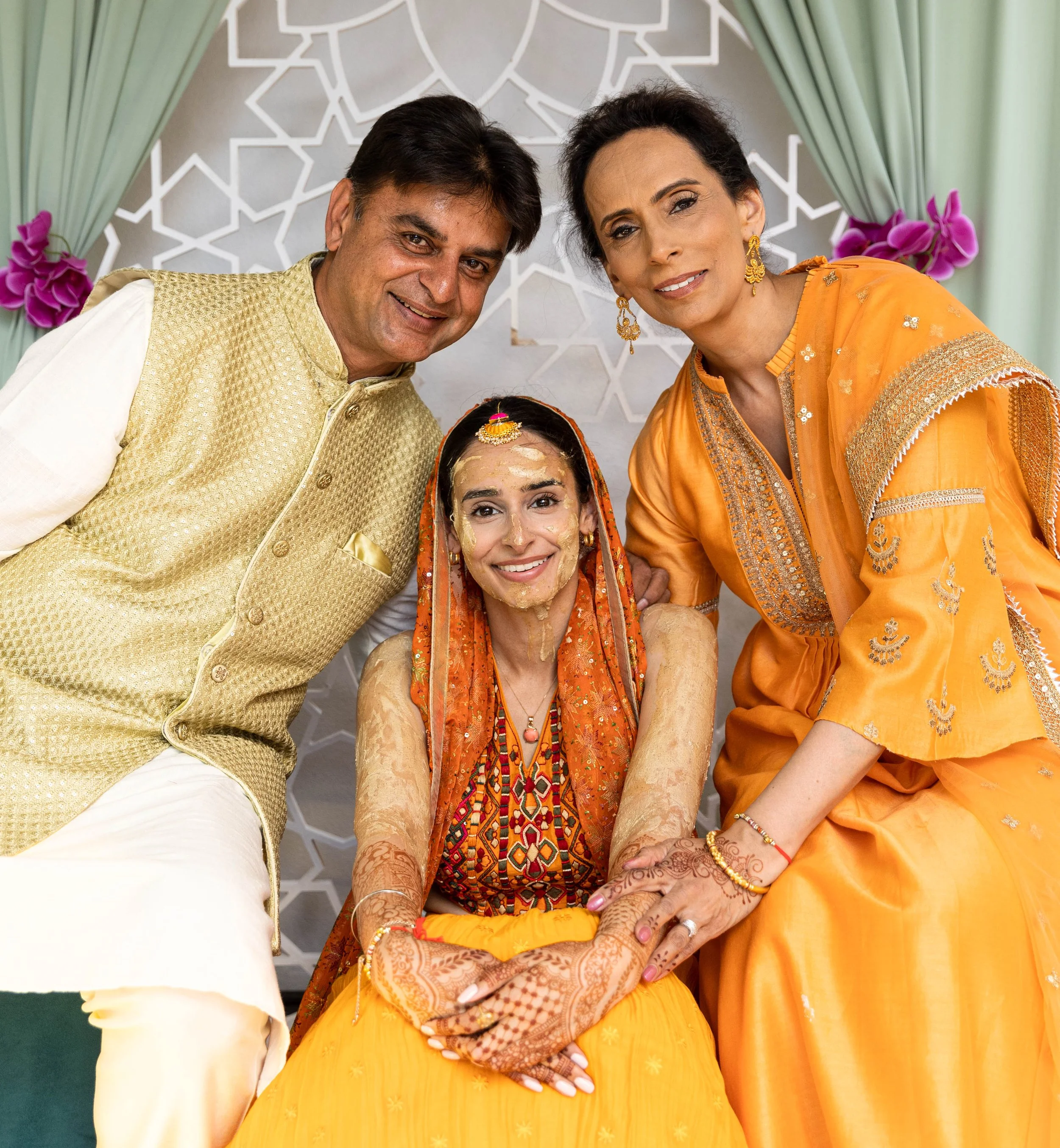 A woman in traditional Indian wedding attire sits smiling, flanked by a man in cream and gold attire on her left and a woman in orange traditional dress on her right, all smiling at the camera.