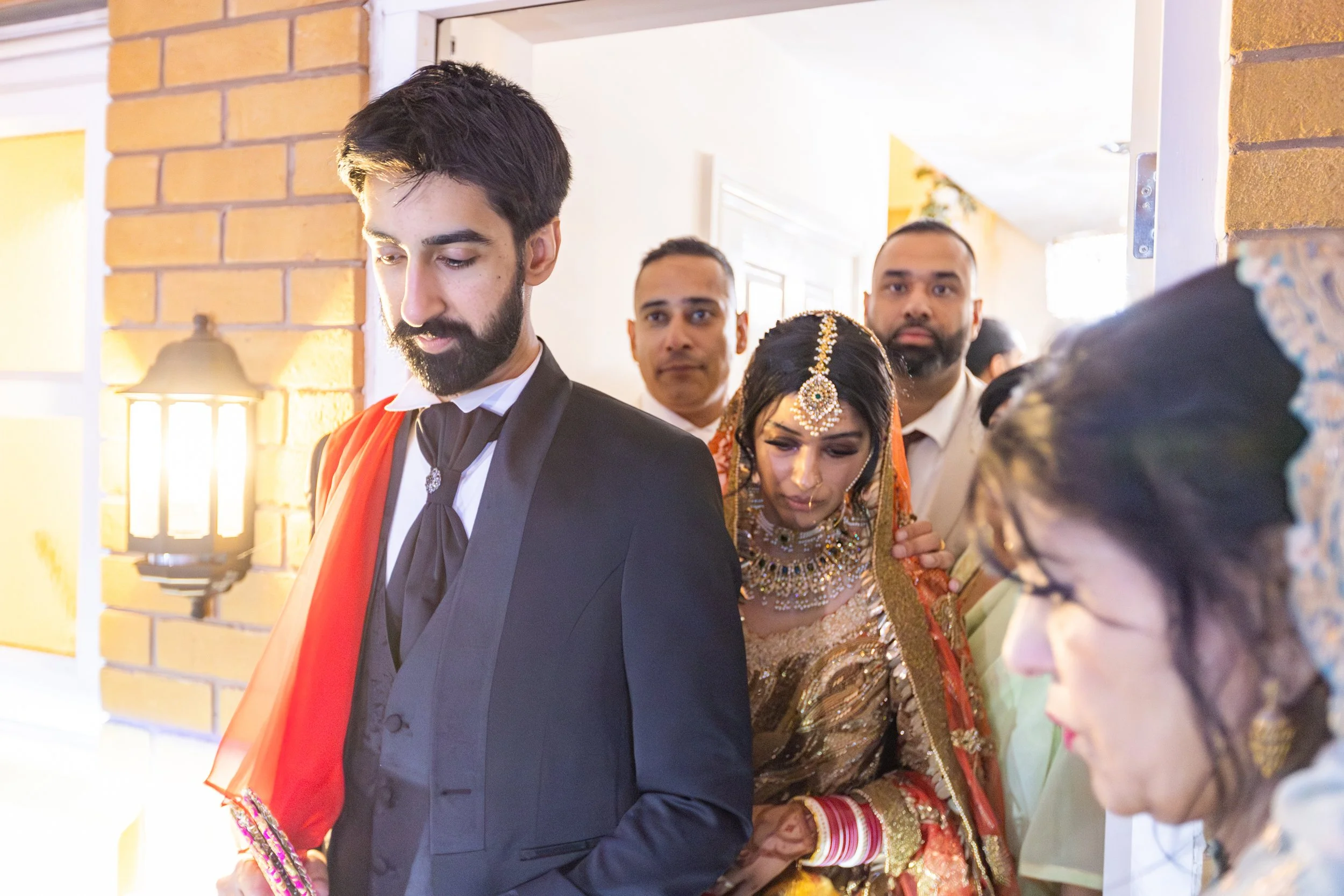A group of people, including a bride in traditional Indian attire and jewelry, and a groom in a suit, gathered indoors during a wedding celebration.