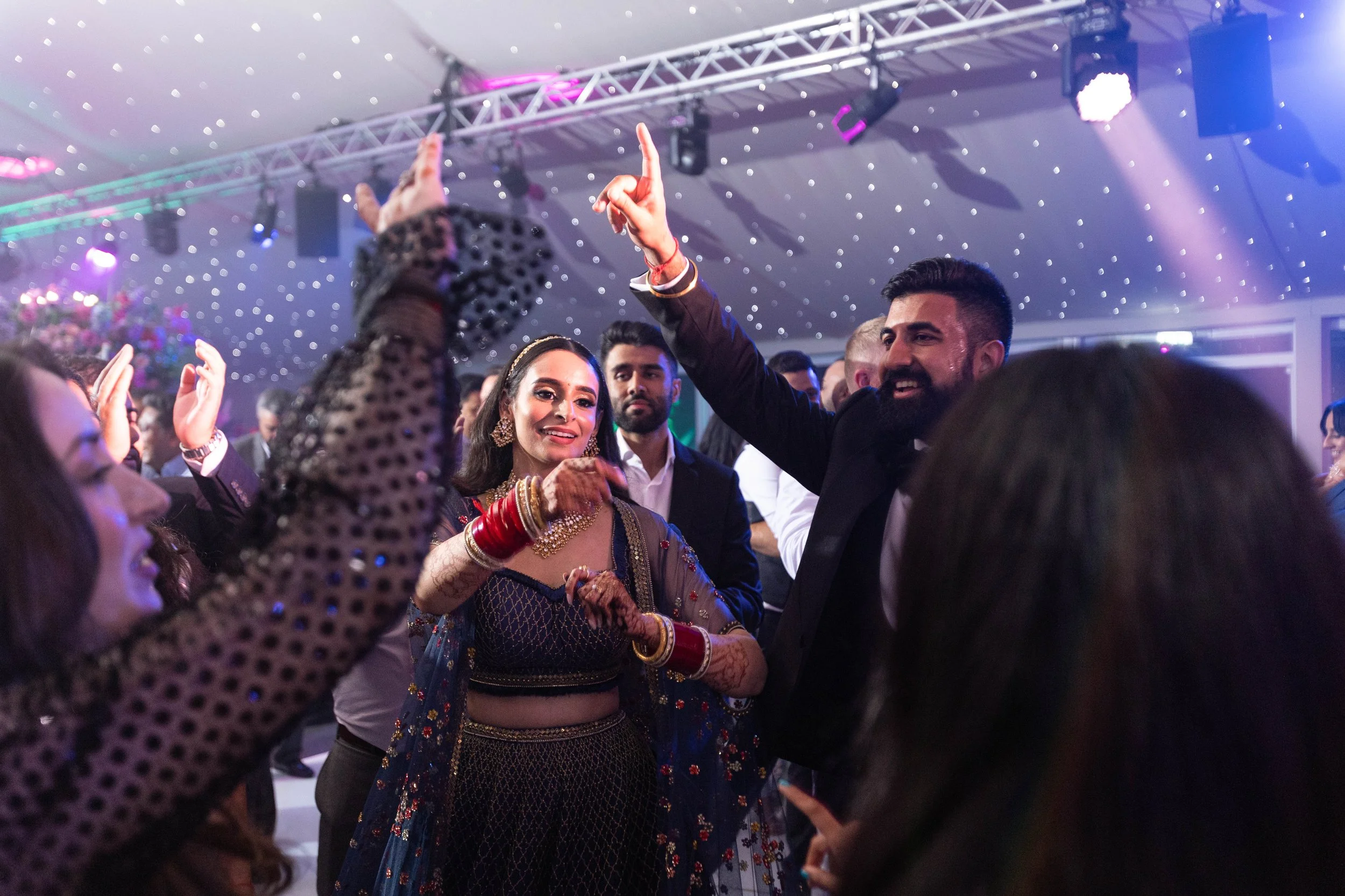 A group of people dancing and celebrating at a wedding reception with colorful lighting and a decorated ceiling.