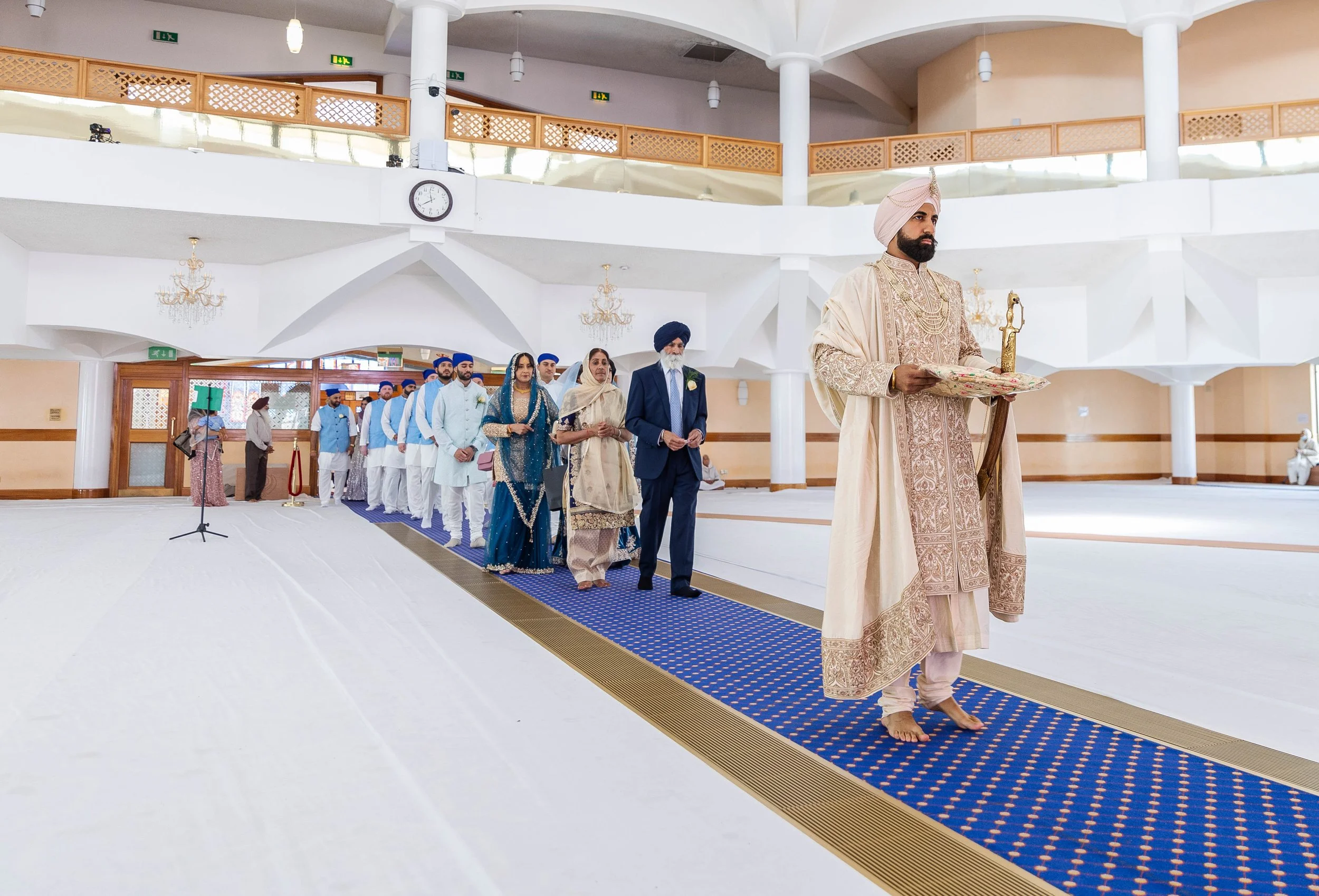 A traditional Sikh wedding ceremony taking place inside a spacious hall. The groom, dressed in ornate traditional Indian attire and a turban, stands at the front holding a tray. The bride and family members, dressed in colorful Indian clothing, are l