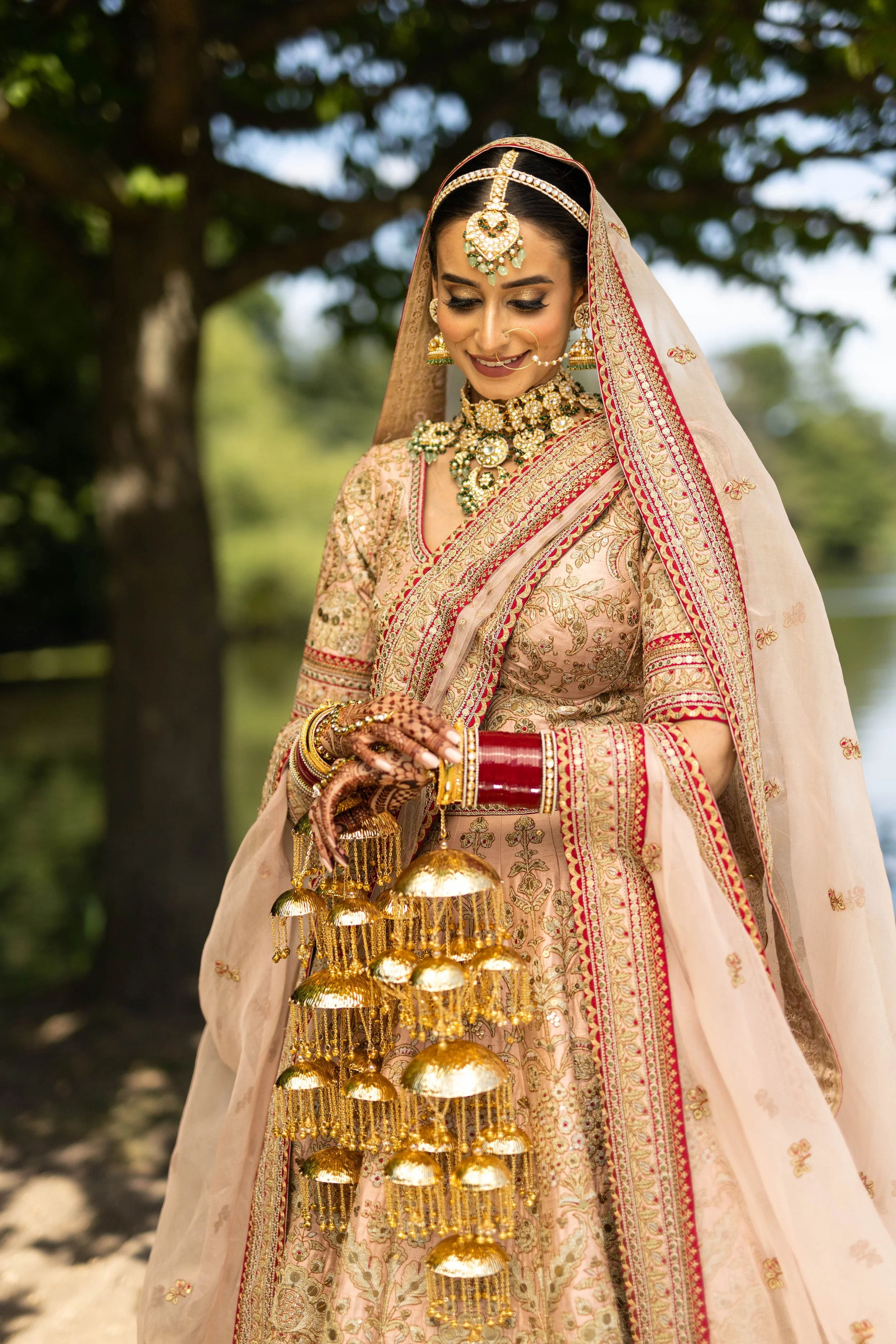 A woman dressed in traditional Indian bridal attire outdoors, wearing a heavily embroidered beige and pink saree, adorned with gold jewelry including a necklace, earrings, forehead ornament, and bangles. She holds a ceremonial tray with hanging gold 