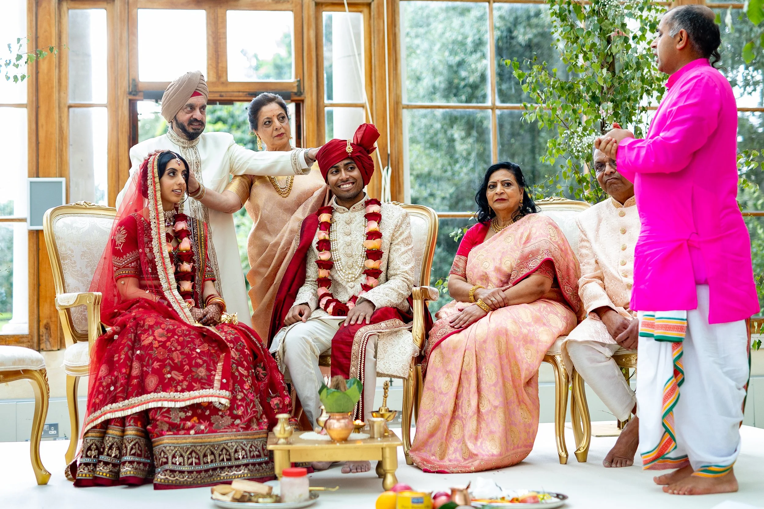 Indian wedding ceremony with bride and groom seated on chairs, surrounded by family members, indoors with large windows and greenery outside, during a traditional ritual.