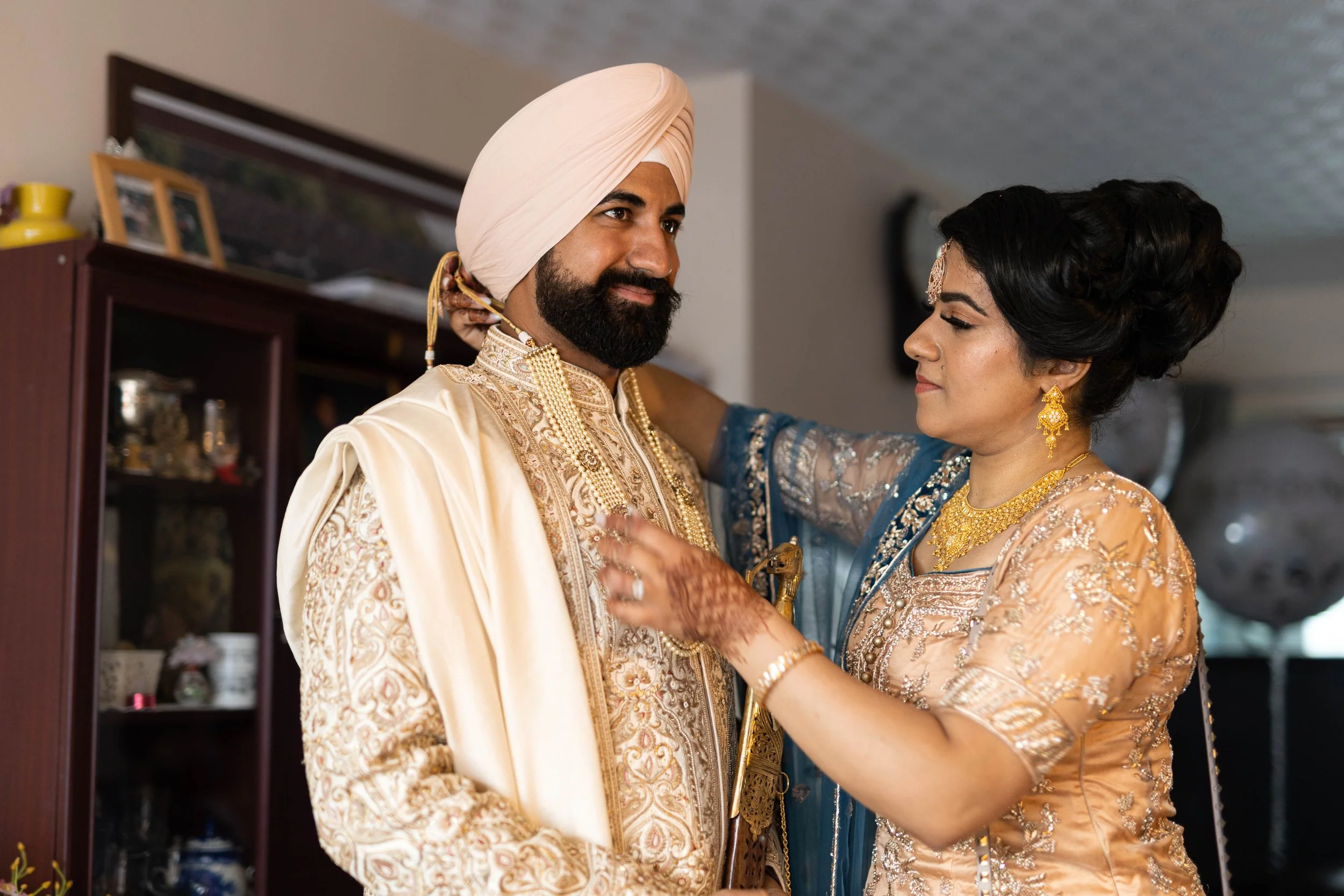 A Sikh groom in cream-colored traditional attire and turban, and a woman in a peach and blue dress, sharing a moment during a wedding ceremony indoors.
