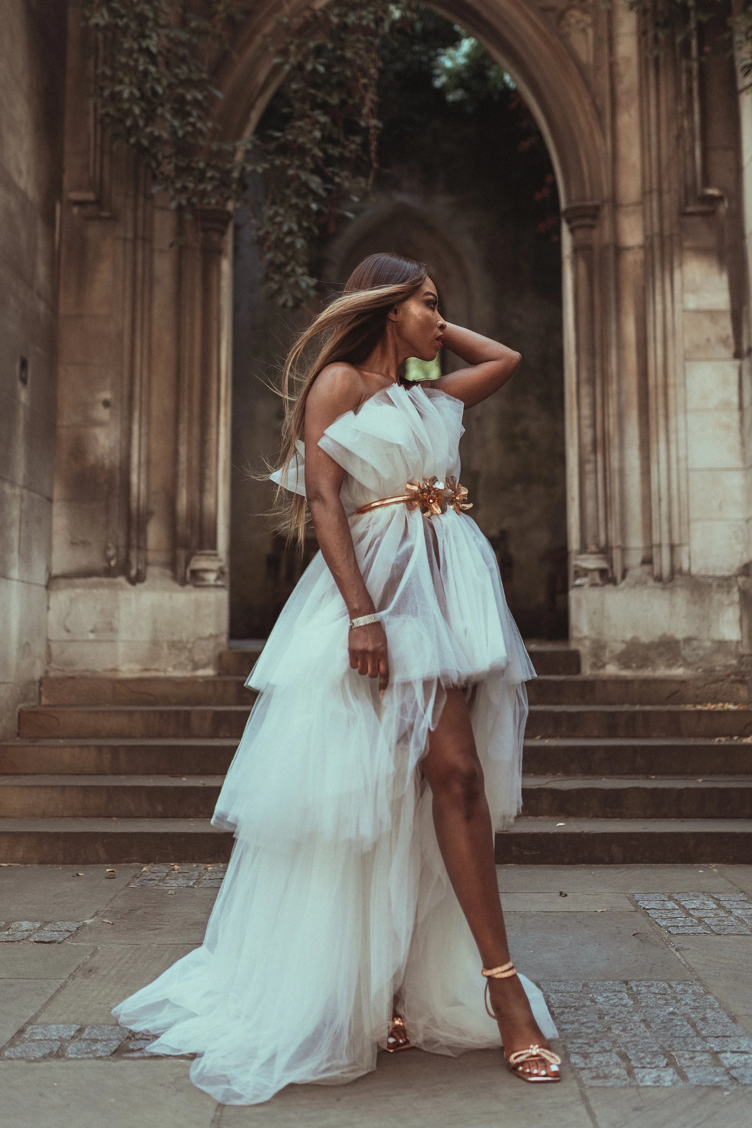 A woman wearing a white tulle dress standing on stone steps outside an old stone building with an arched doorway, surrounded by greenery.