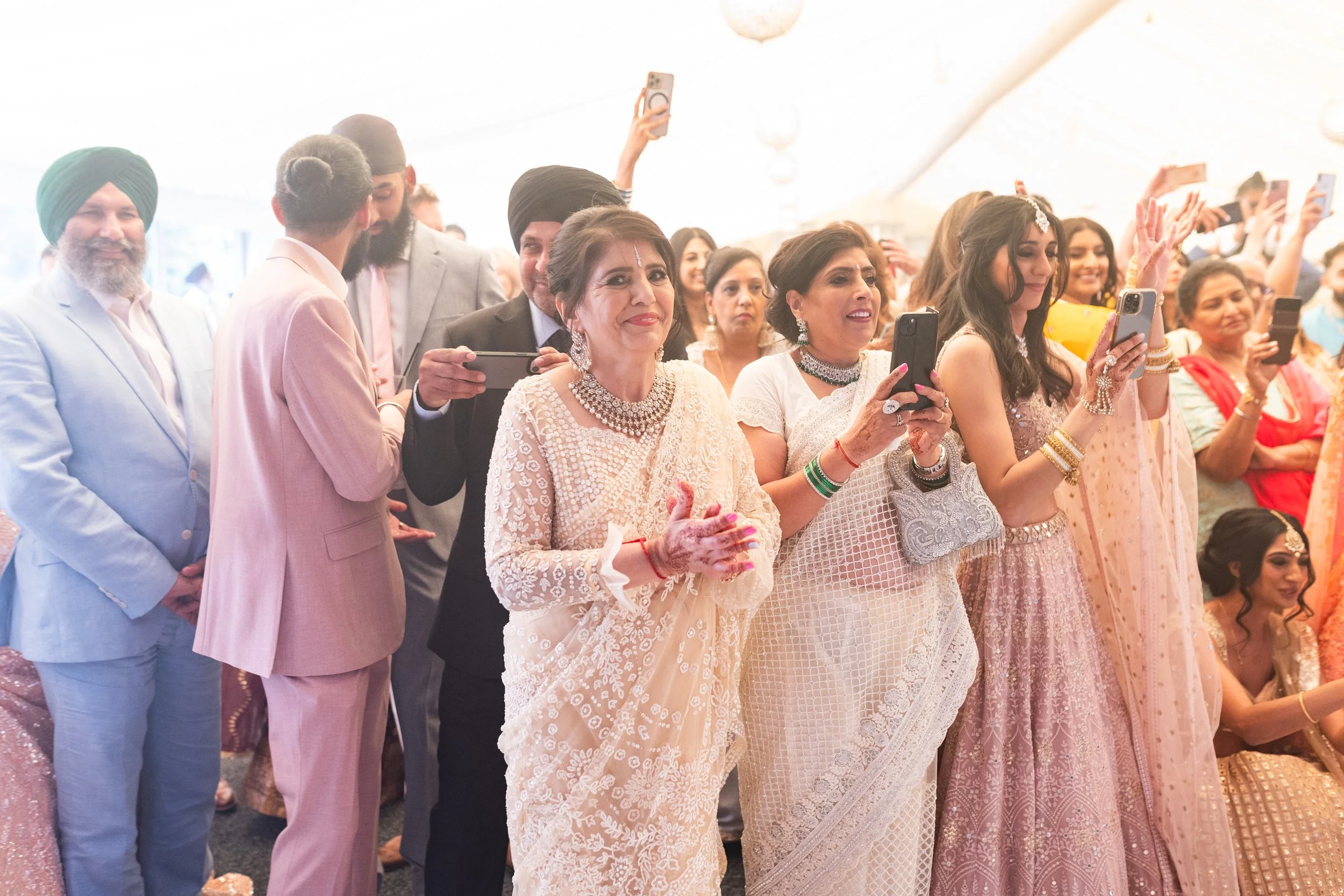 Group of people attending a celebration, many dressed in traditional Indian attire, some holding smartphones to take photos.