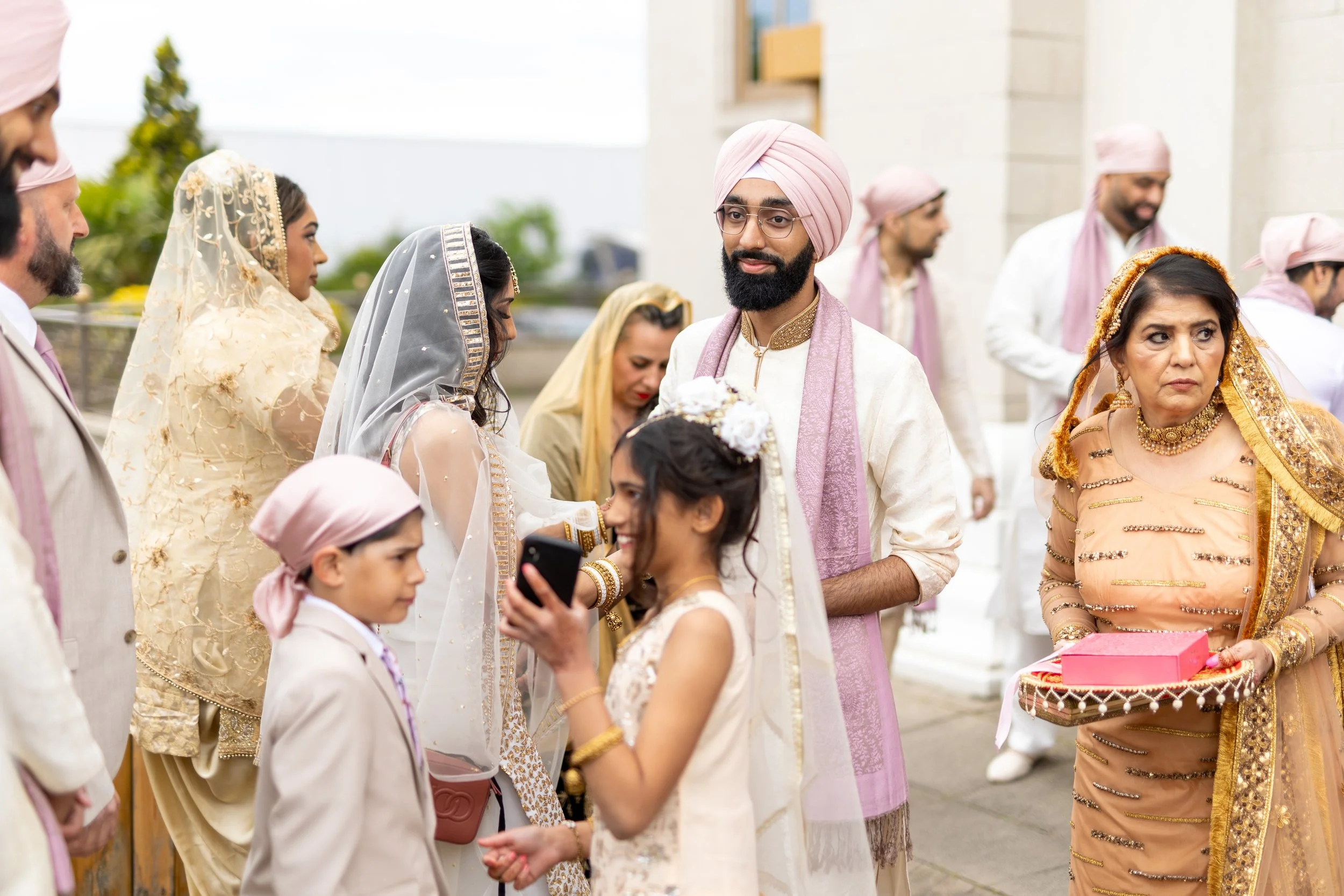 Indian wedding ceremony with guests dressed in traditional attire, women in sarees and men in kurtas, outdoors on a cloudy day with buildings and greenery in the background.
