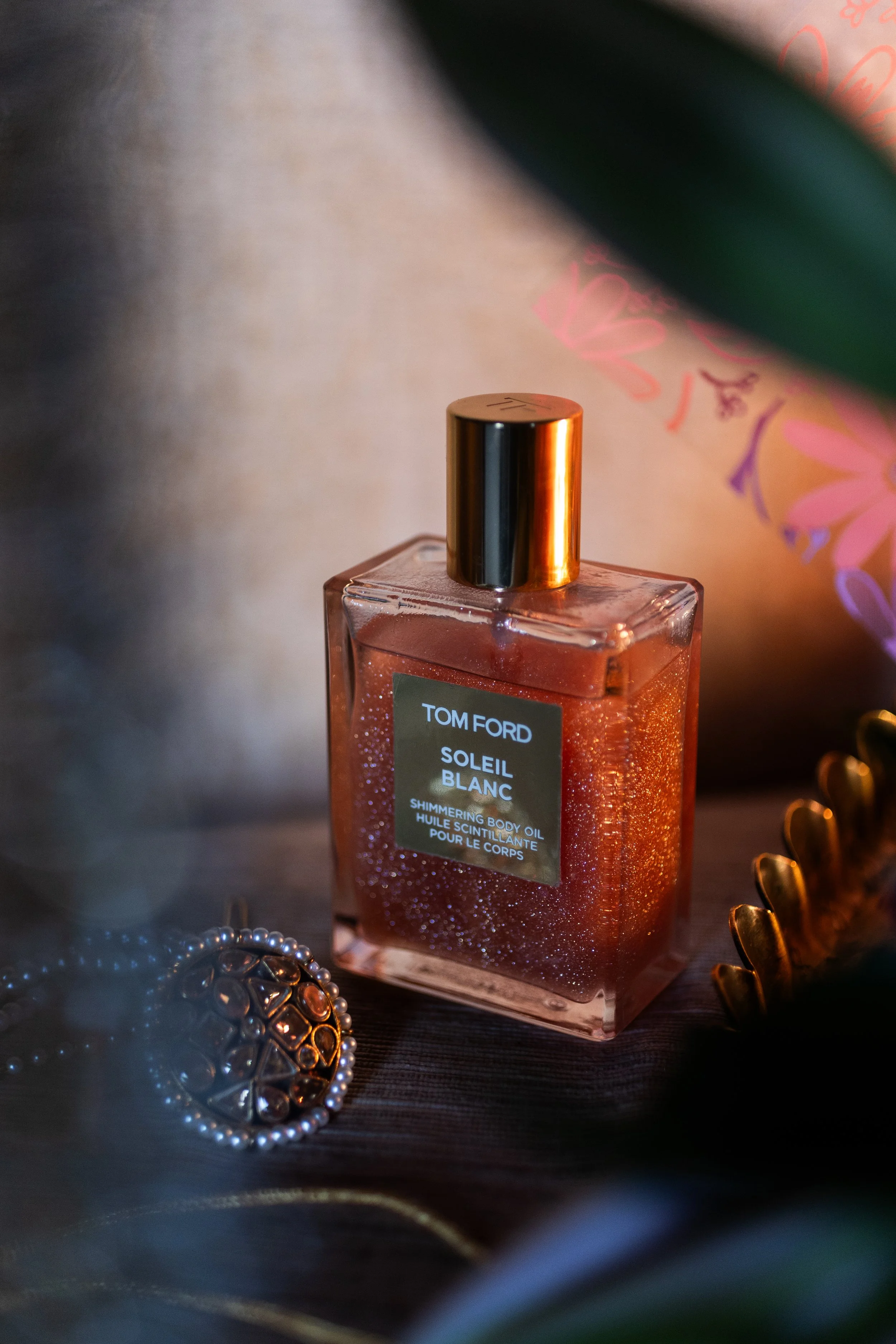 A bottle of Tom Ford Soleil Blanc shimmer body oil with a gold cap, placed on a dark surface with jewelry and a floral-patterned background.
