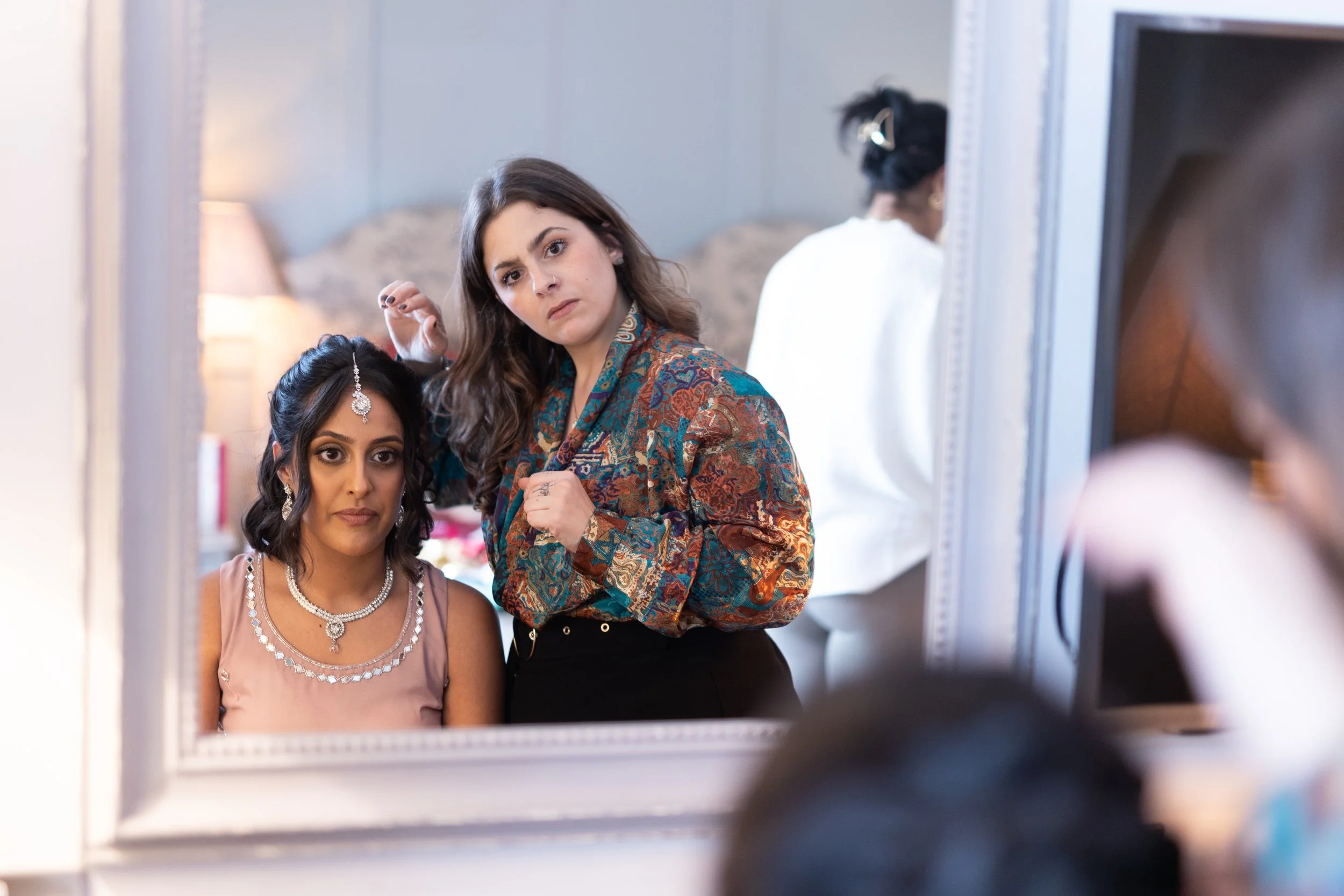 Woman with jewelry sitting in front of mirror, another woman with long hair and patterned blouse fixing hair, both looking into a mirror, with another person in the background