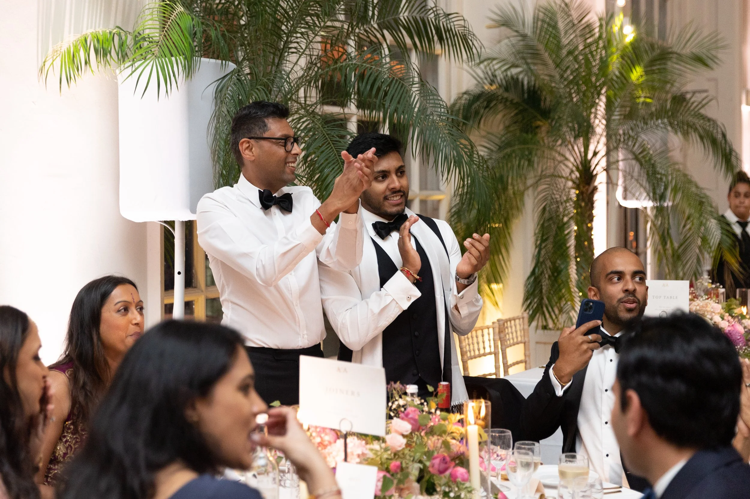 Group of people at a formal event, with two men standing and clapping, one wearing glasses and both in tuxedos with bow ties, surrounded by seated guests at a decorated table with flowers and candles, lush greenery background.