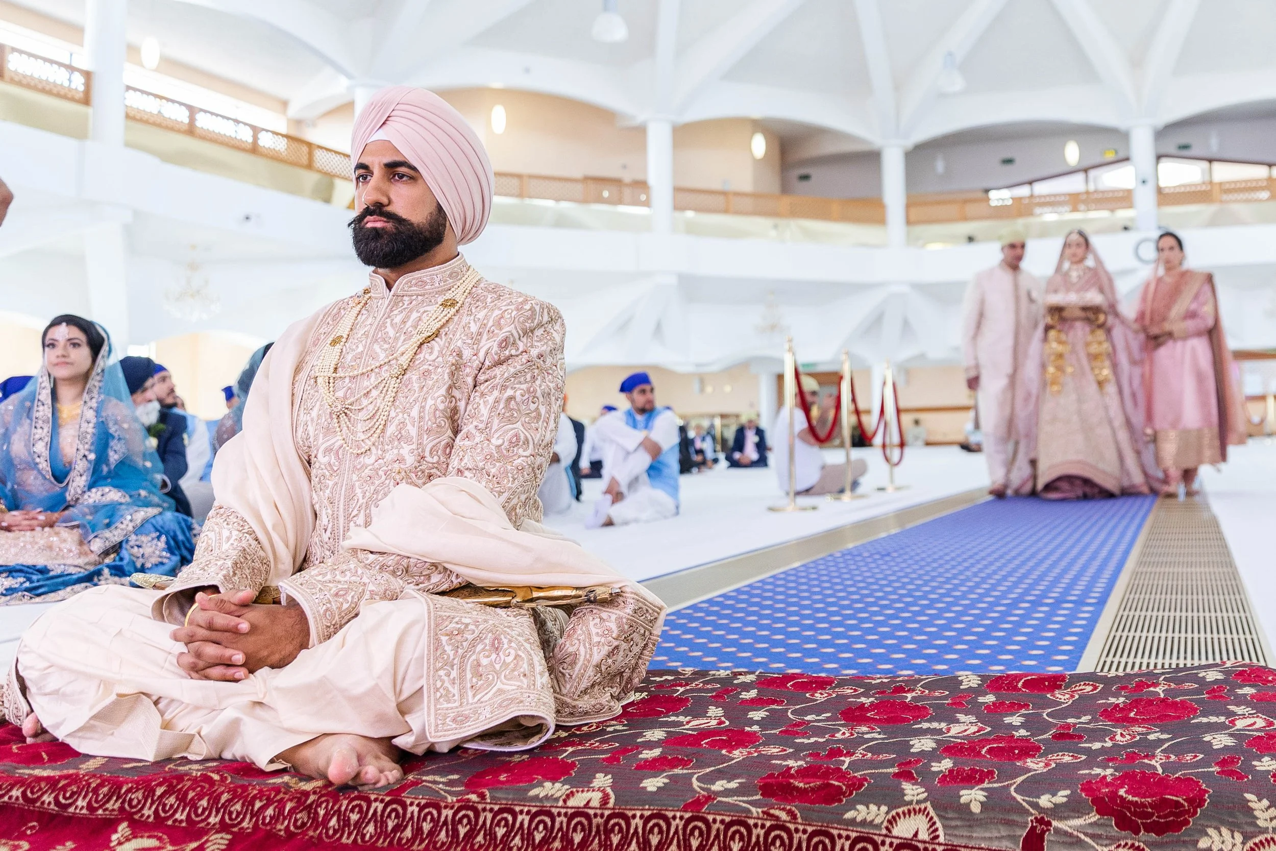 A man wearing traditional Indian groom attire, seated on a carpet during a wedding ceremony. Other people dressed in traditional attire are visible in the background inside a spacious hall.