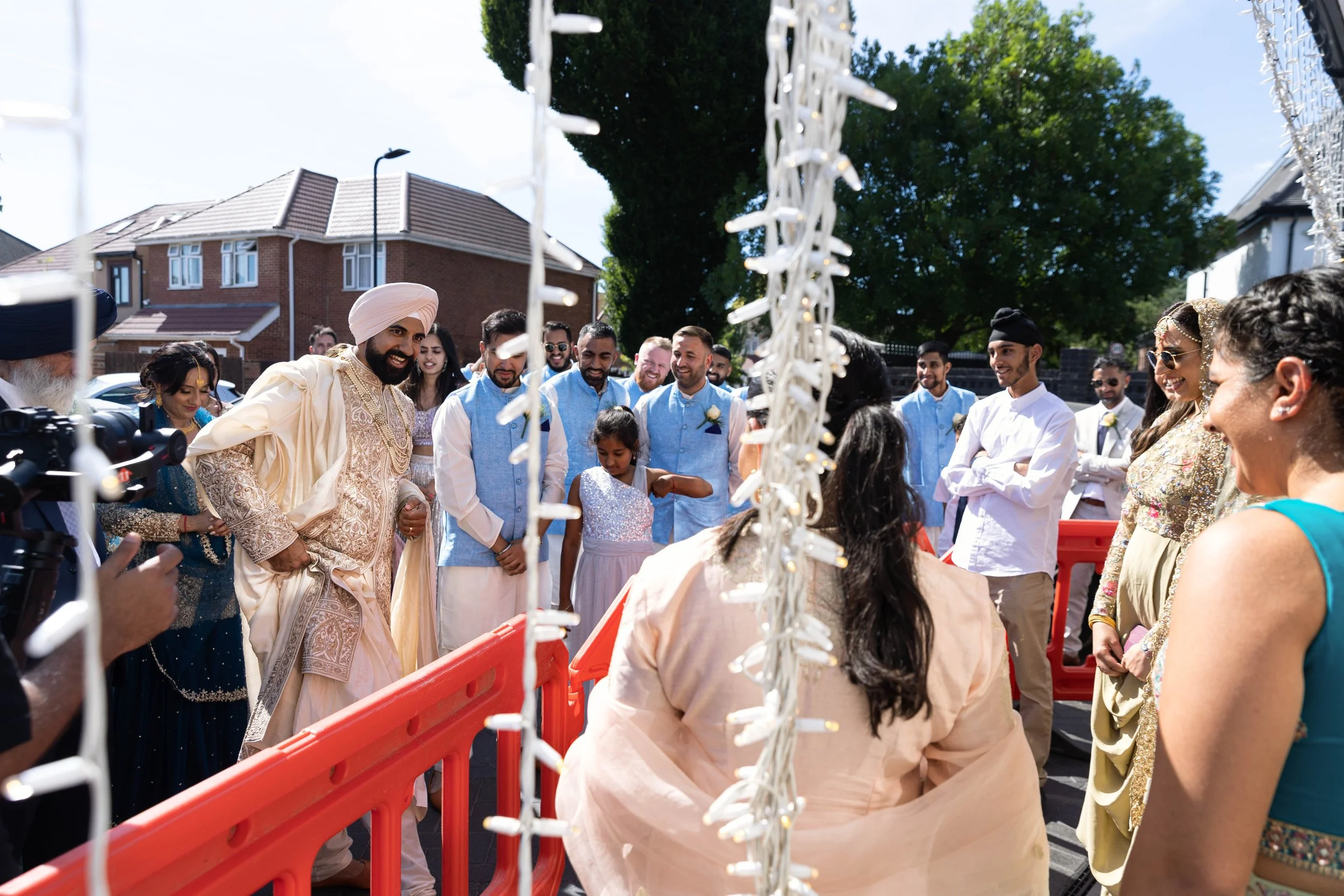 Indian wedding celebration outdoors with people dressed in traditional attire, smiling and socializing near a decorated area with string lights, in a residential neighborhood.
