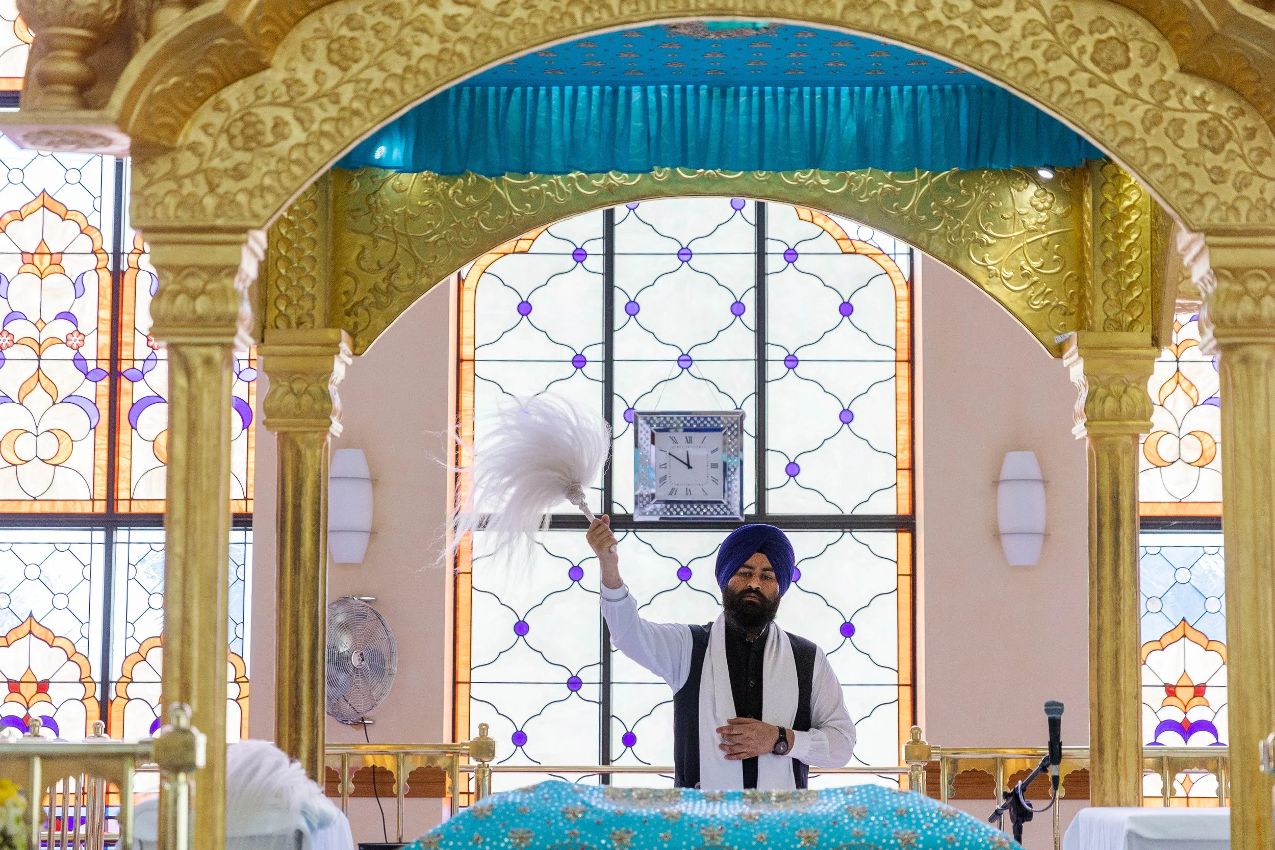 A man dressed in traditional Sikh attire, including a blue turban, performing a religious ritual inside a decorated prayer hall with stained glass windows and ornate golden arches. He is holding a white feathered ceremonial item.