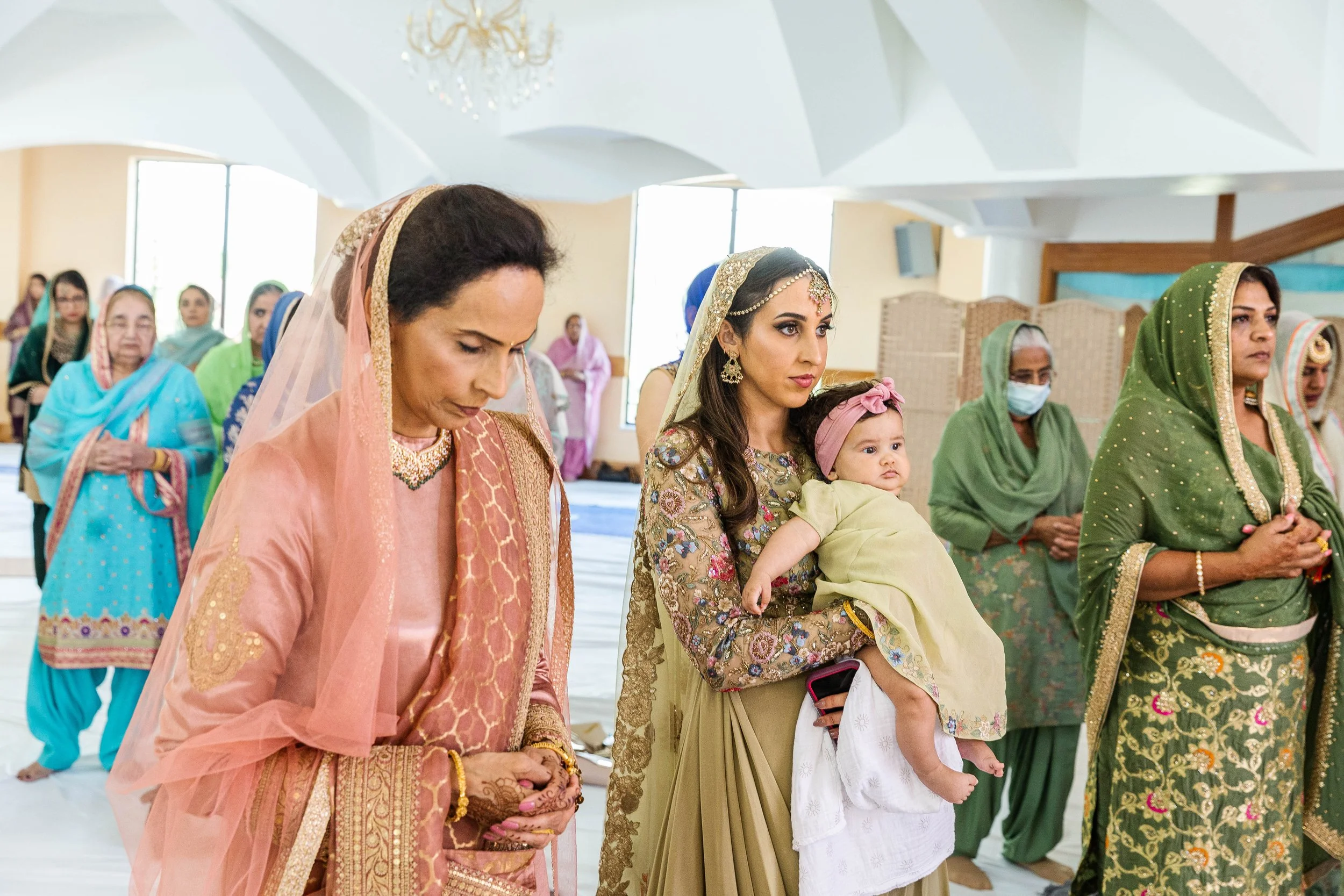 Women and a baby participating in a religious or cultural ceremony, with women wearing colorful traditional Indian attire and praying with their hands folded in a temple setting.