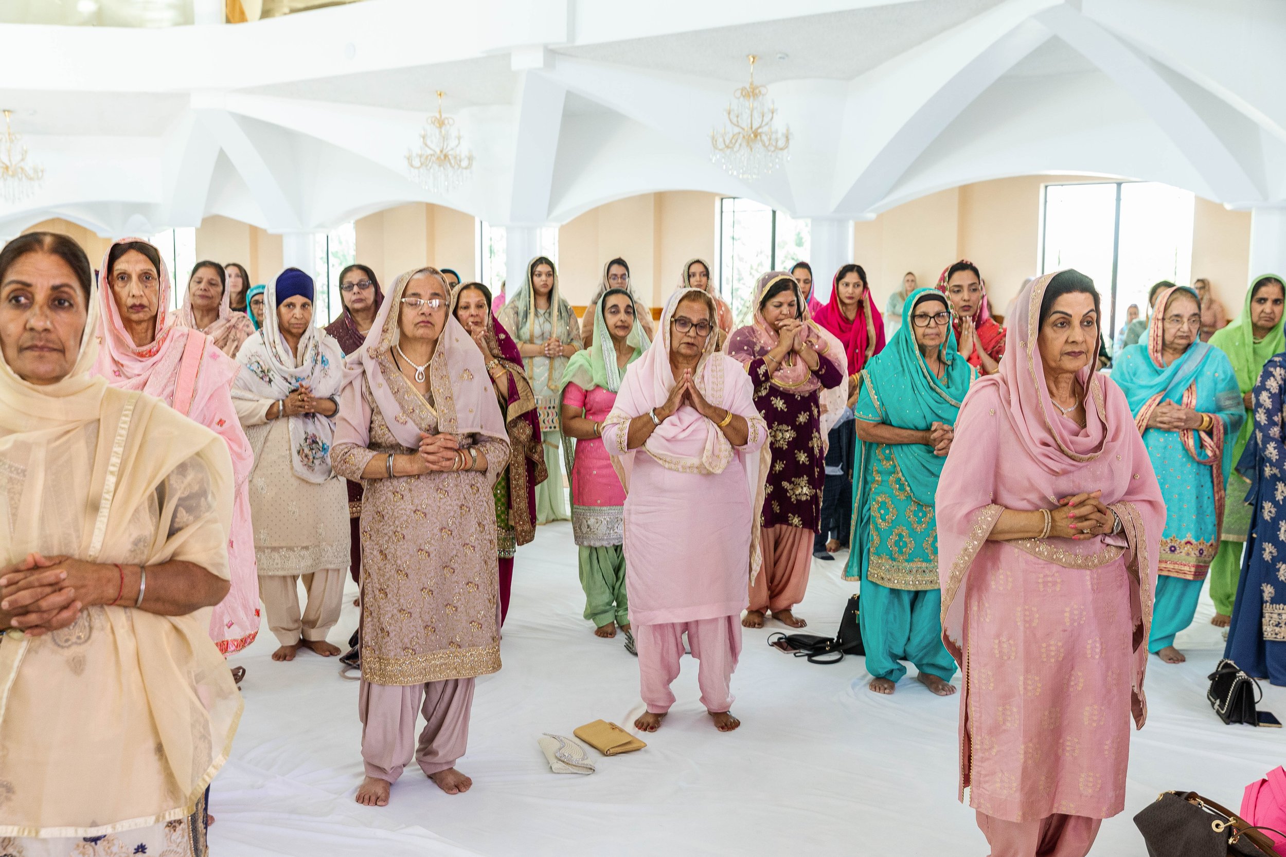 Group of women in colorful traditional Indian dresses participating in a prayer or religious ceremony inside a church with white walls, chandeliers, and large windows.