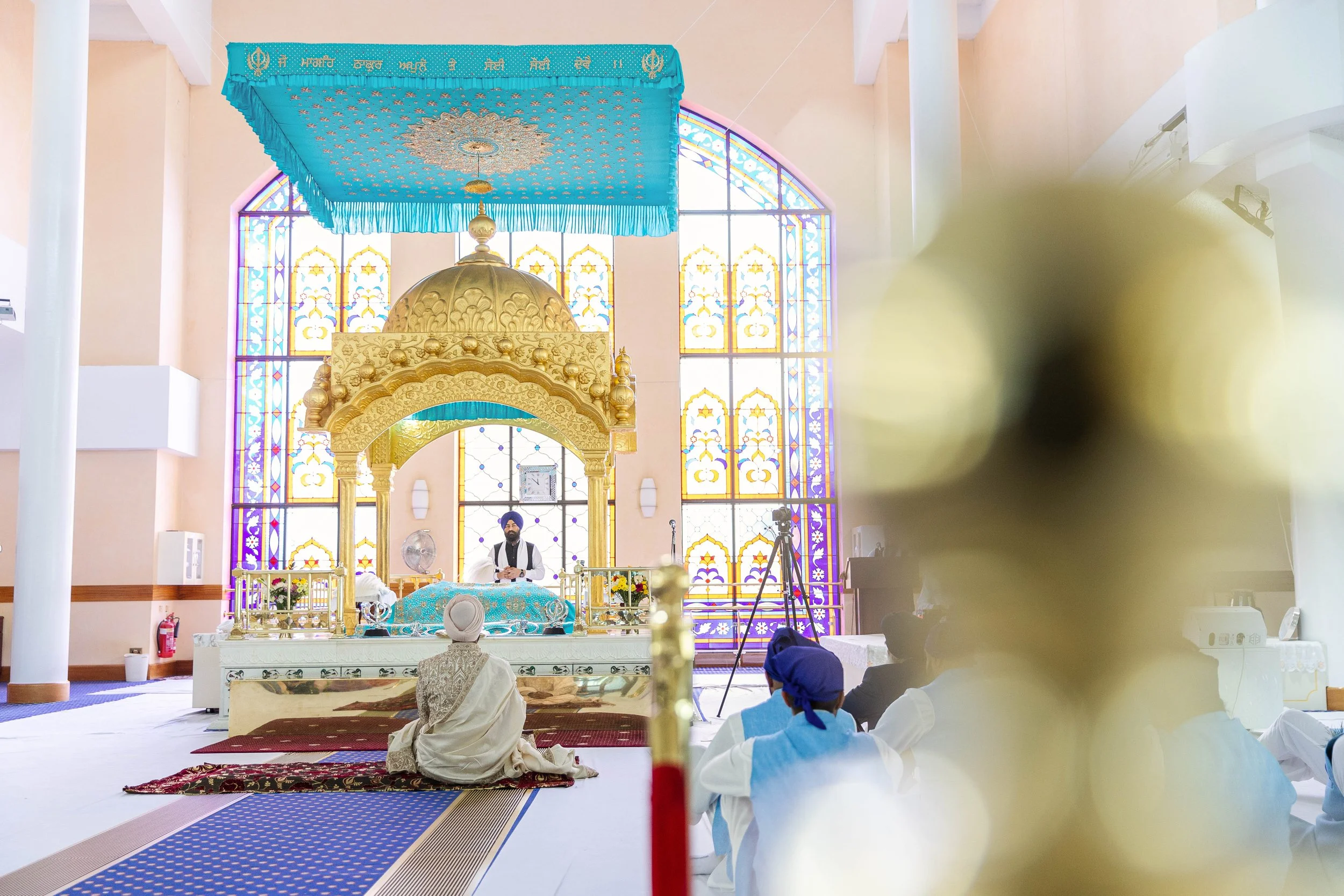 A Sikh wedding ceremony takes place inside a gurdwara with stained glass windows, a sacred canopy, and a person sitting at the front in traditional attire. There are other attendees seated on the floor, with a floral decoration in the foreground.