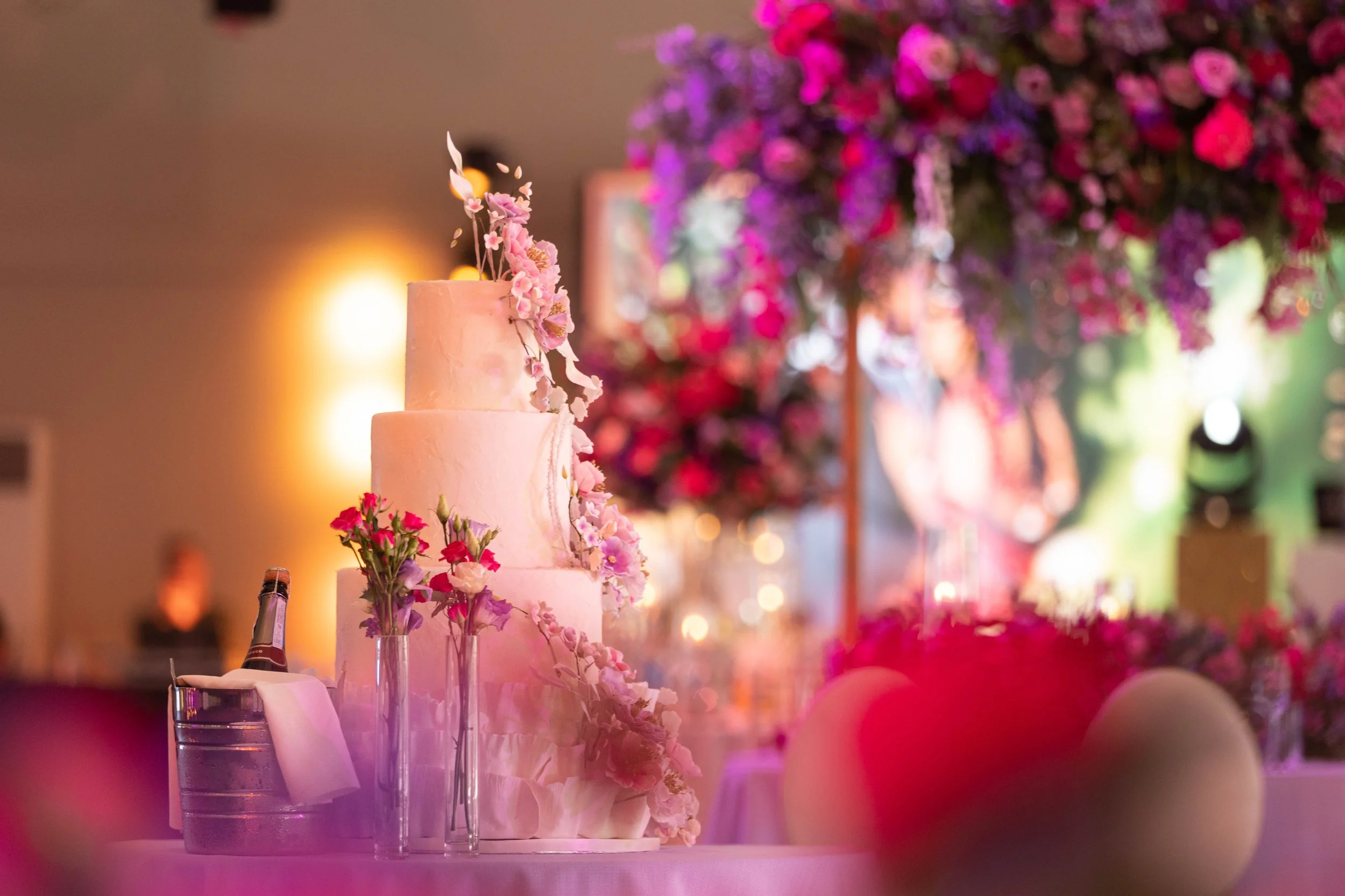 A multi-tiered wedding cake decorated with pink and purple flowers, placed on a table with small flower vases and a bottle in an elegant reception hall with pink, purple, and green floral arrangements