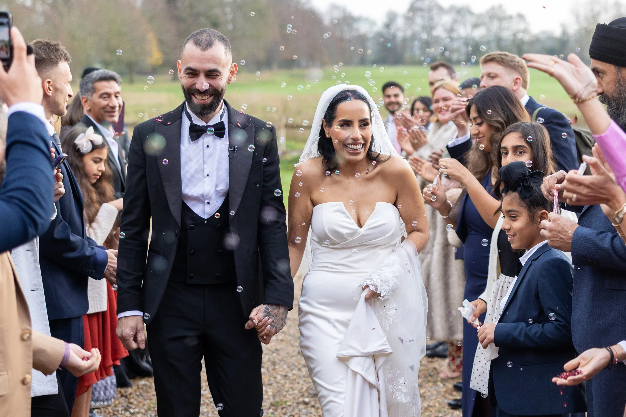 Happy newlywed couple holding hands and walking through a crowd of wedding guests outdoors, surrounded by bubbles, on their wedding day.